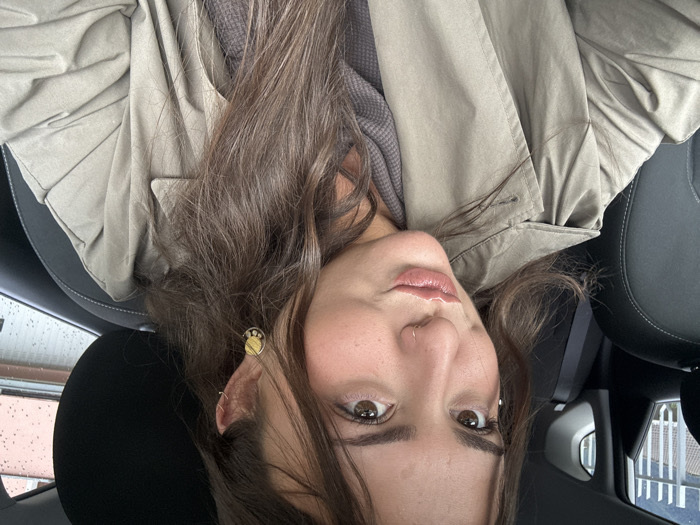 A young adult female is centered, posing for a selfie from the front passenger seat of a vehicle. She has long, wavy, dark brown hair, brown eyes, and a puckered lip expression with light, glossy lipstick. A small silver hoop piercing is present in her right nostril, and a similar hoop is in her left helix. A circular gold-toned dangle earring with a patterned design adorns her right earlobe. She wears a light olive-green trench coat over a gray, textured long-sleeved garment. The vehicle's interior shows black synthetic or leather upholstery. Raindrops are visible on the exterior side window. The out-of-focus background reveals a light-colored building facade with dark horizontal lines and a metal railing structure. This scene is located in the city of Nesso, Italy.