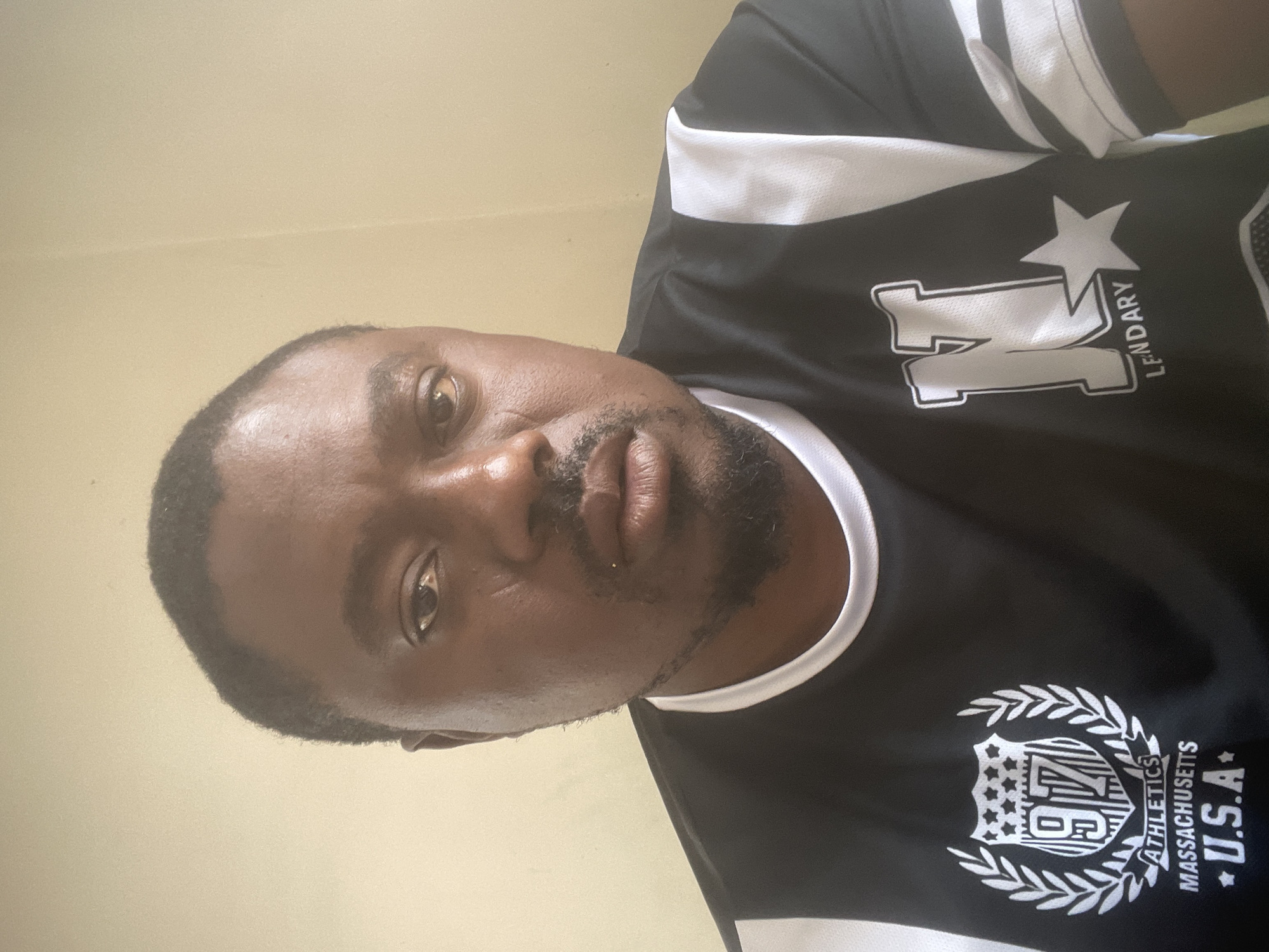 The media file displays a self-portrait of an adult man, likely taken indoors in Jos, Nigeria. The image is rotated 90 degrees clockwise, showing the man's head tilted towards the left, suggesting the photograph was taken with the camera held sideways. He has a dark complexion, short dark hair, and a short beard or stubble. He is looking directly at the camera with a neutral expression.
The man is wearing a black short-sleeved t-shirt with white accents, including white stripes on the sleeves and white trim around the collar. The shirt features several white printed designs. On his left side (viewer's right), there is a large stylized "N" symbol with a star above it, and the word "LENDARY" printed vertically beside the "N." On his right side (viewer's left), there is a crest-like emblem. This emblem contains the number "97" within a shield, surrounded by a laurel wreath. Below the shield, the text "ATHLETICS MASSACHUSETTS U.S.A." is clearly visible, with a star next to "U.S.A.".
The background consists of a plain, light-colored wall, likely beige or cream, indicating an indoor setting. There are no other visible objects or distinct features in the background. The lighting appears even and sufficient, suggesting daytime conditions or well-lit indoor environment. The man's activity is posing for the camera.