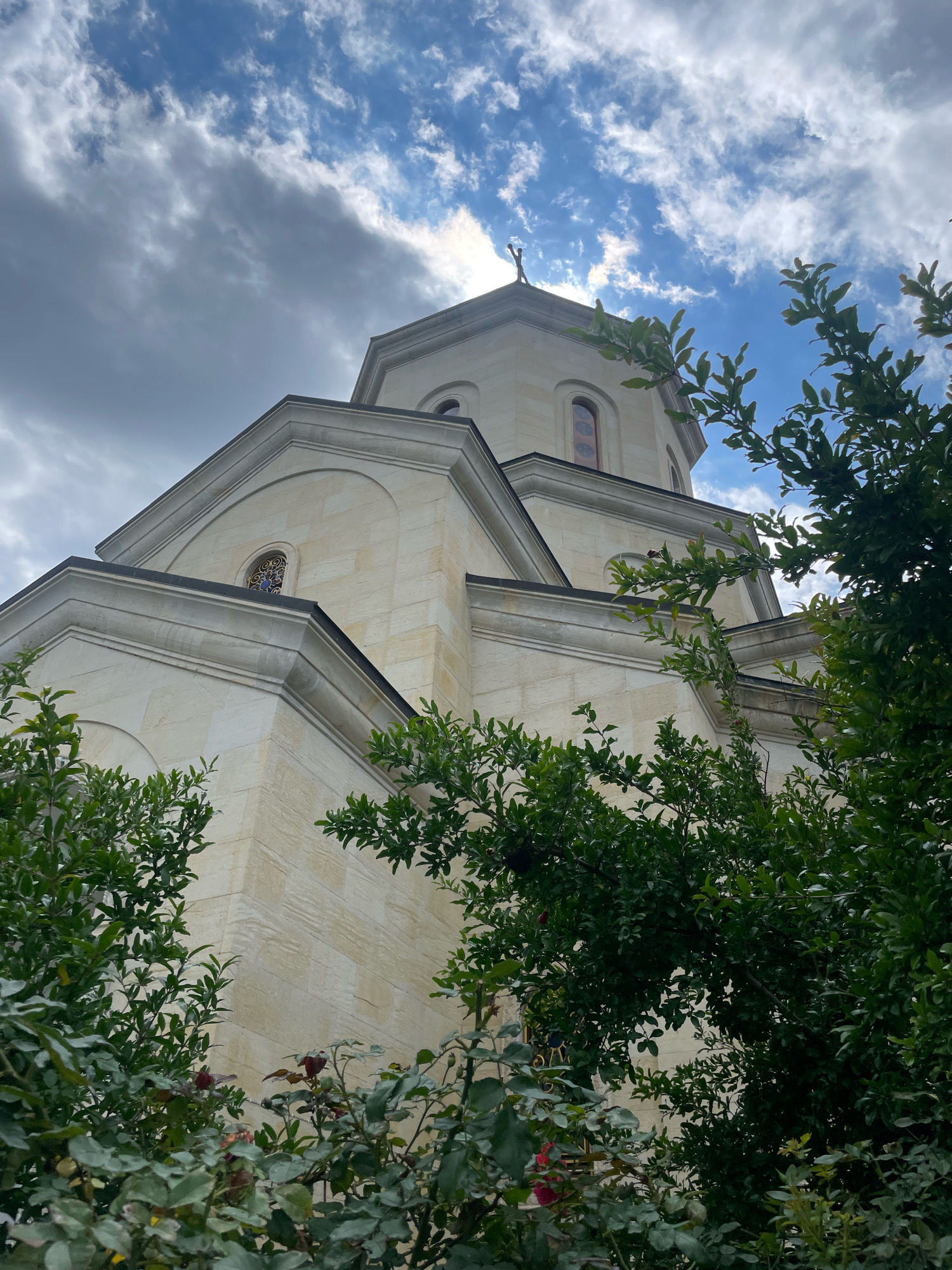 The image shows the exterior of a pale stone church, possibly a chapel or a small cathedral, against a partially cloudy blue sky. The church has a domed roof topped with a cross. The architecture features clean lines, arched windows, and decorative trim. Lush green foliage, including trees and roses with some red buds, partially obscures the lower portion of the building, framing it from the foreground. The scene is captured from a low angle, looking upwards at the structure. This church is identified as the Church of Saint George in Saguramo, Georgia.