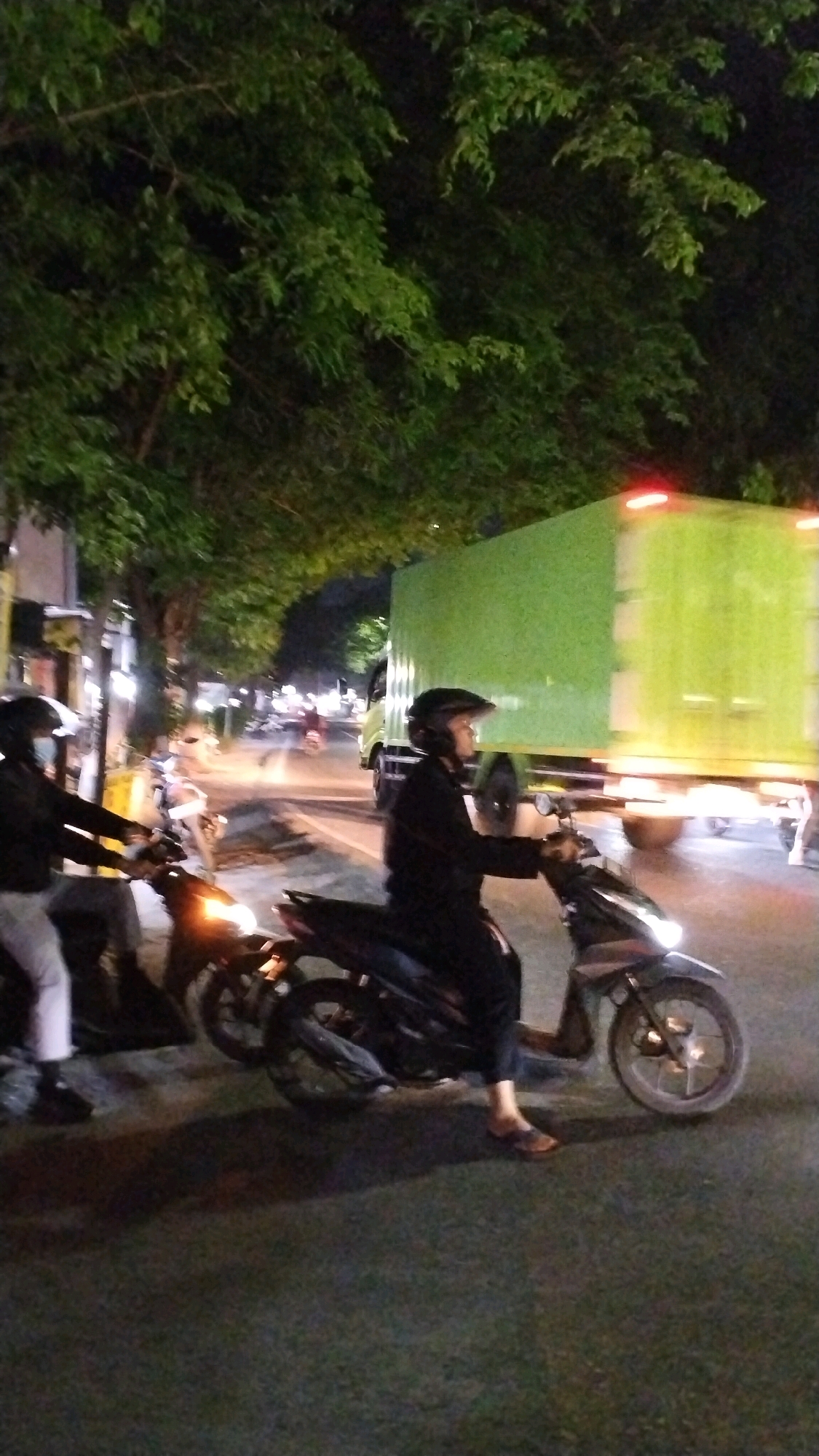 In Canggu, Indonesia, a street scene unfolds at dusk or night, with lush green foliage overhead partially obscuring the view of the sky. A large, bright green box truck is a prominent feature, its rear lights illuminated. In the foreground, two individuals are on motorcycles. The person on the right wears a black jacket and helmet, with their right hand extended forward, seemingly operating the motorcycle. Their left leg is bent at the knee, and they are wearing sandals. The motorcycle they are riding is dark colored, with a visible headlight and wheel. To the left, another person in a black jacket and helmet is partially visible, also on a motorcycle. This motorcycle also has its headlight illuminated, and the rider appears to be wearing light-colored pants and dark shoes. The road surface is visible, and there are other vehicles and lights in the background, suggesting traffic.