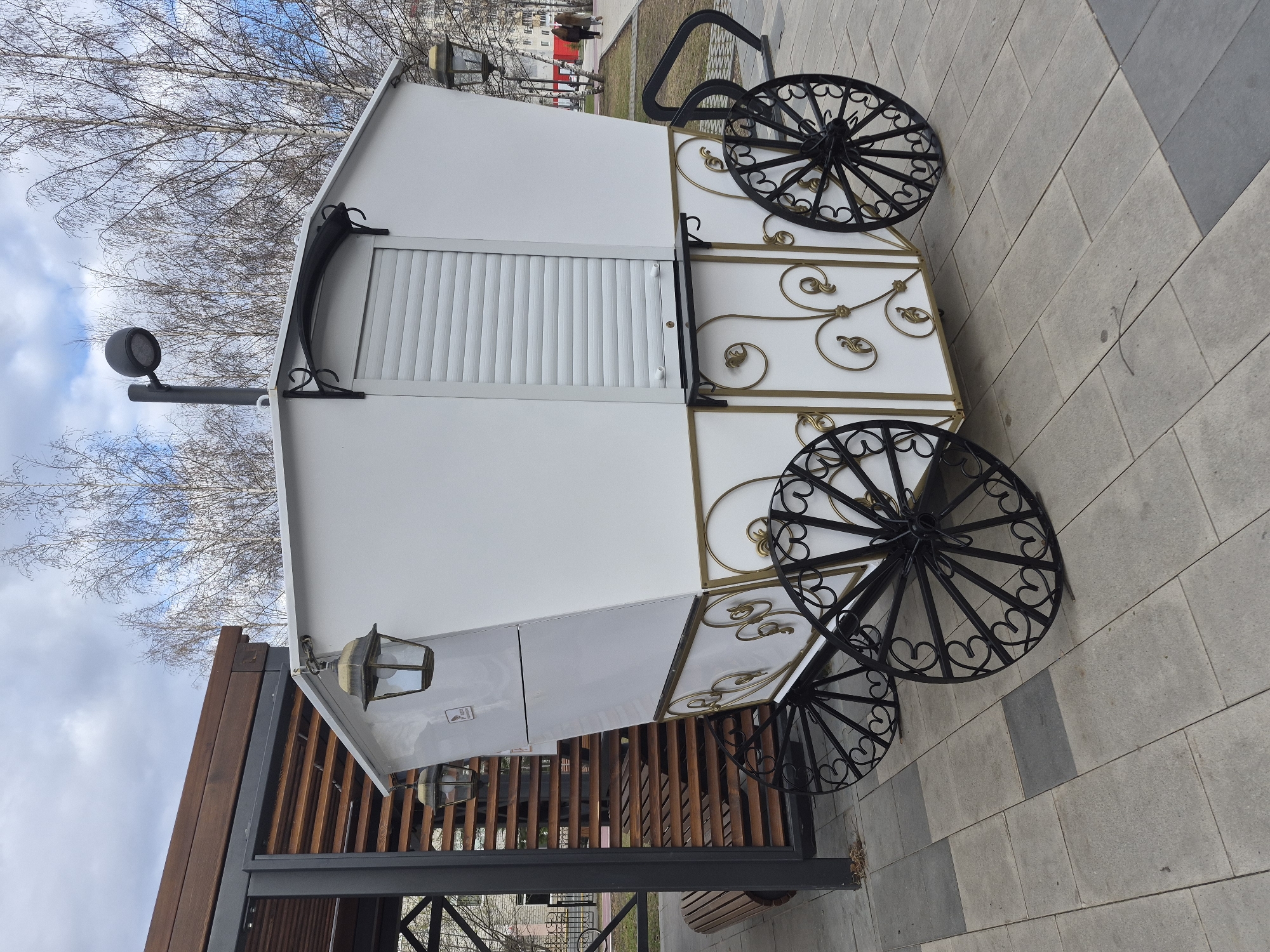 A white, carriage-style kiosk or small building with ornamental black wrought iron wheels and gold filigree trim sits on a gray brick-paved area. The structure has a closed white rolling shutter, a black dome light on a pole, and a vintage-style lamp. The kiosk is near a wooden structure with vertical slats, a black metal frame, and a view of a park-like area with trees and benches. Other people are in the distant background, walking and standing. The sky is blue with patchy clouds, and bare trees suggest it might be late fall or winter. This is located in the city of Tobolsk, Russia.