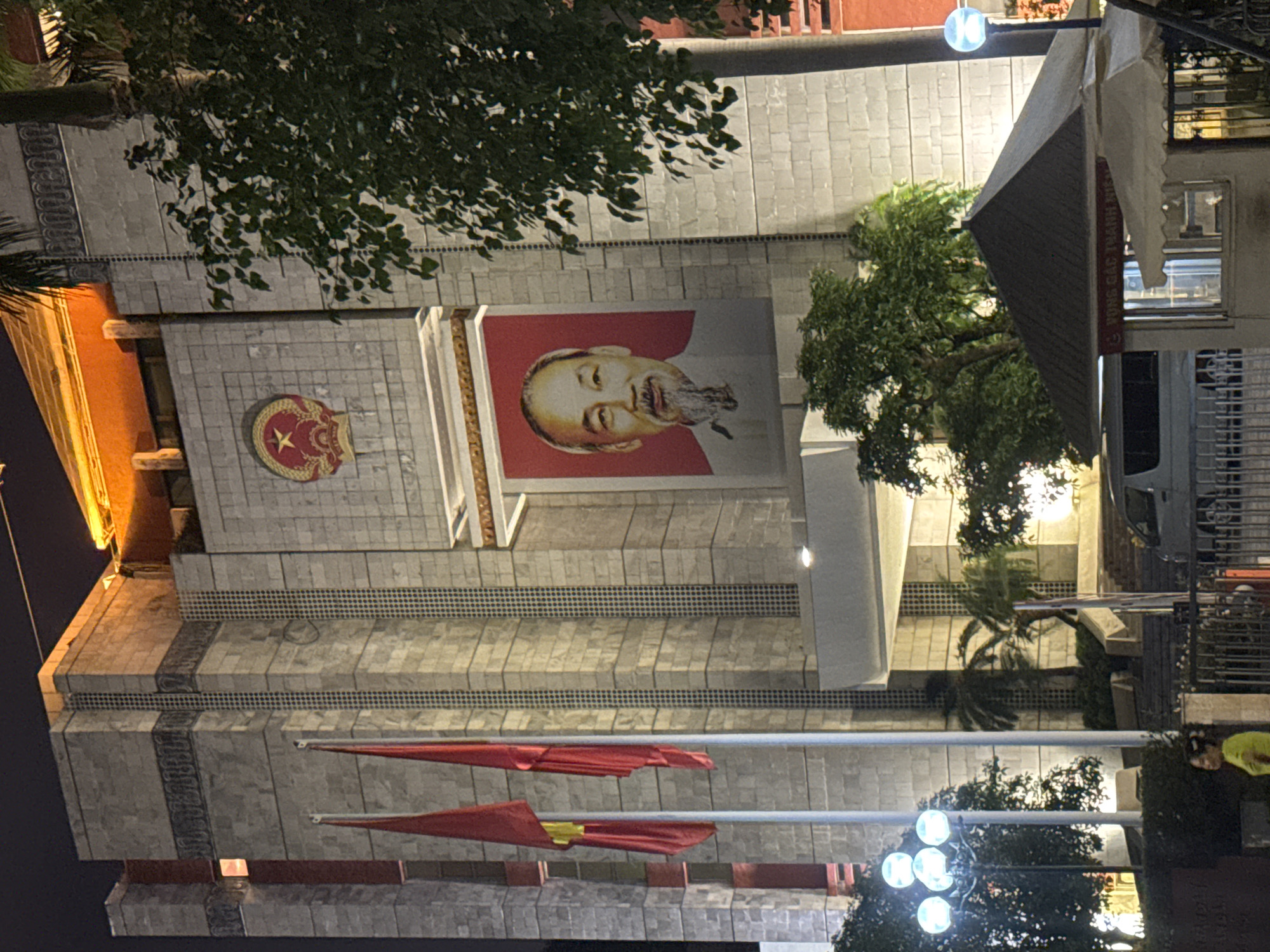 The image shows the Hồ Chí Minh Mausoleum in Hà Nội, Vietnam at night. A large portrait of Ho Chi Minh is displayed prominently on the mausoleum's facade, along with the Vietnamese flag. Several red flags hang in front of the building. Streetlights illuminate the area, and trees partially obscure the view of the building. A street-side store is located along the right of the picture with some parked vehicles nearby. A person wearing a green vest is also visible on the right side. The Mausoleum is constructed of grey stone.