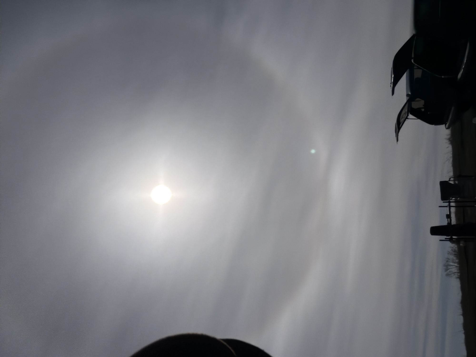 The image shows a bright sun in the sky with a visible halo surrounding it. A structure appears on the right side of the image. The environment consists of an overcast sky. The photograph is taken from an angle, making it difficult to determine the objects and features in the image. The image does not show signs that it was taken in Nikolo-Berezovka, Russia.