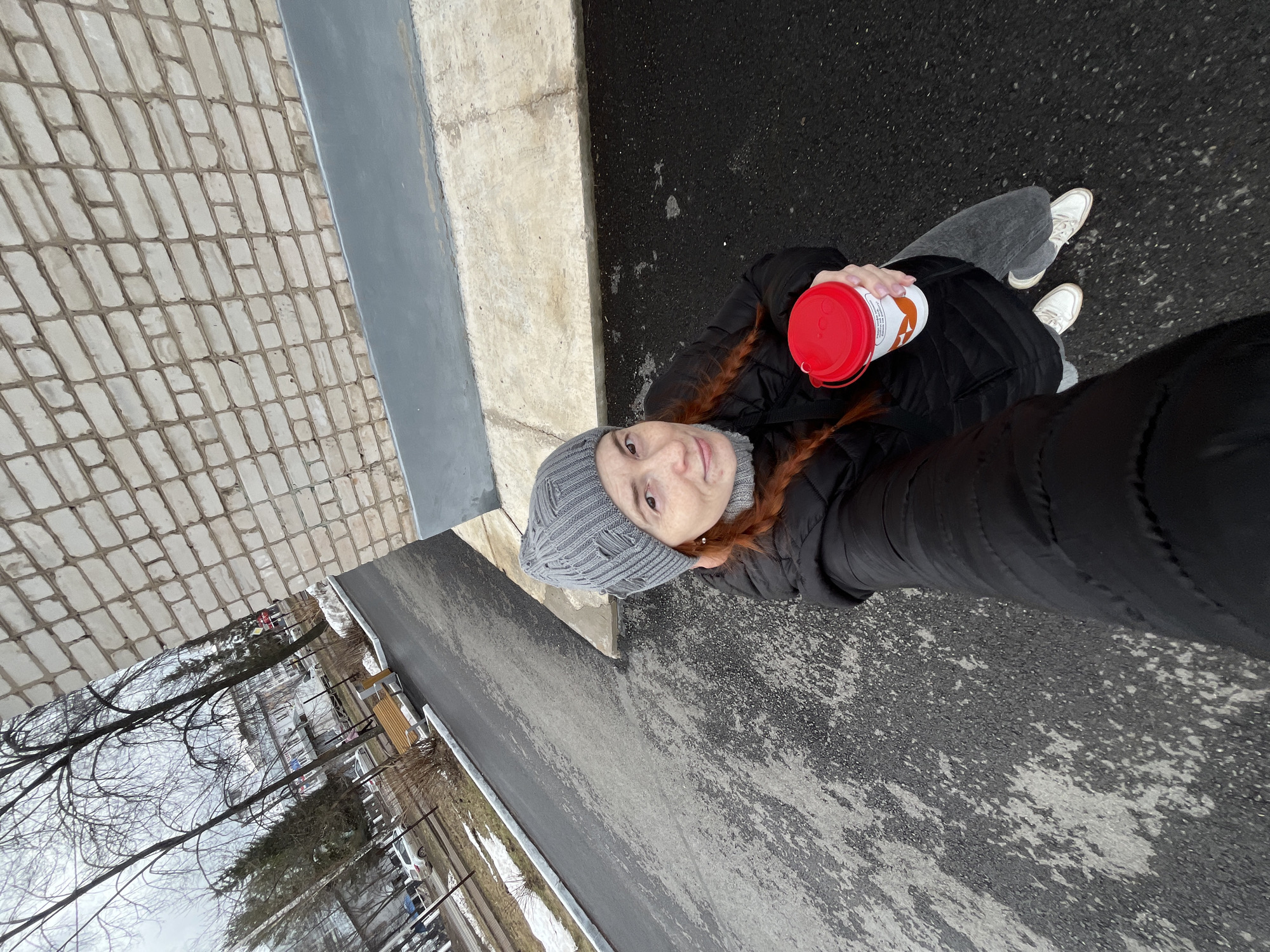 A high-angle view of a woman standing on a pavement or road next to a brick sidewalk and a concrete curb. She is holding a red and white cylindrical container. She's wearing a dark winter coat, gray jeans, white sneakers, and a gray knitted hat. In the background is a residential area in what could be the city of Chaikovsky, Russia, with bare trees and snow patches visible. The perspective suggests the photo was taken with a selfie-style camera angle.