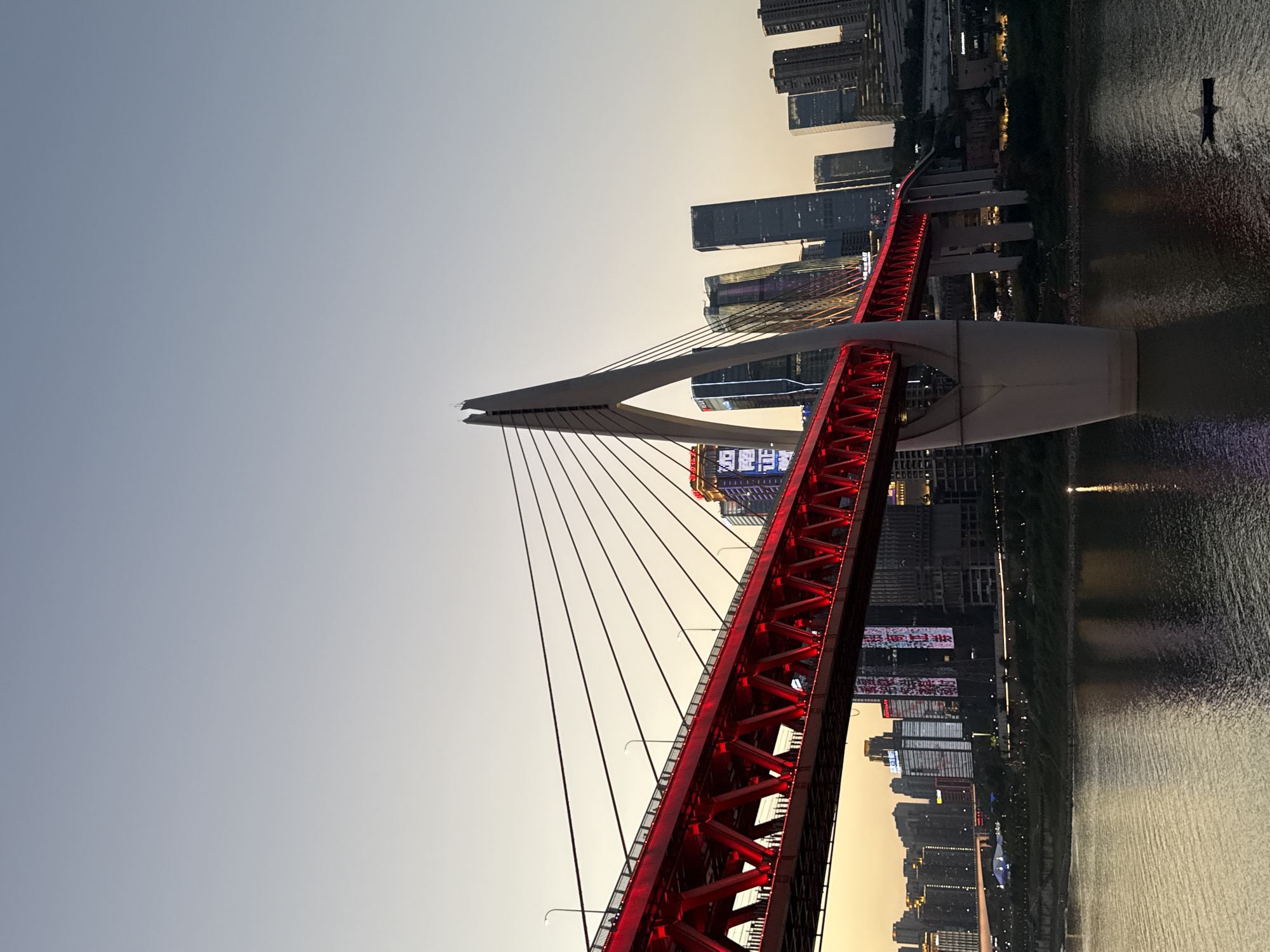The image shows a view of the Hongyadong Bridge in Chongqing, China, during dusk. The bridge, illuminated in red, spans across a body of water. A white pylon supports the suspension cables of the bridge, and the city skyline with illuminated skyscrapers can be seen in the background. The sky is a soft gradient of blue and orange, indicating twilight. The water reflects the lights of the city and the bridge. There is a small boat visible on the water. The scene depicts an urban landscape with modern architecture and infrastructure.