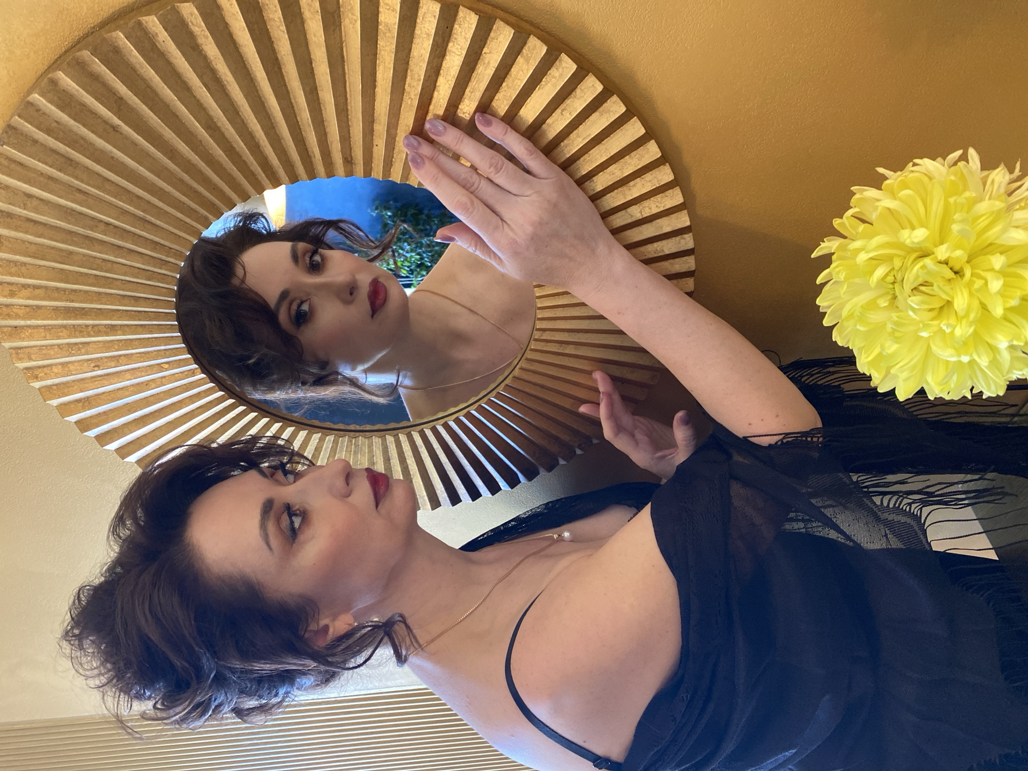 A woman is looking at her reflection in a round, gold-colored mirror with radiating stripes. Her dark hair is styled in waves. She is wearing a black shawl and a gold necklace. A large, yellow chrysanthemum is to the right of the mirror. The background wall is painted in a warm, tan color. The location is not discernible from the image. It is not possible to determine if this is in Yekaterinburg, Russia.