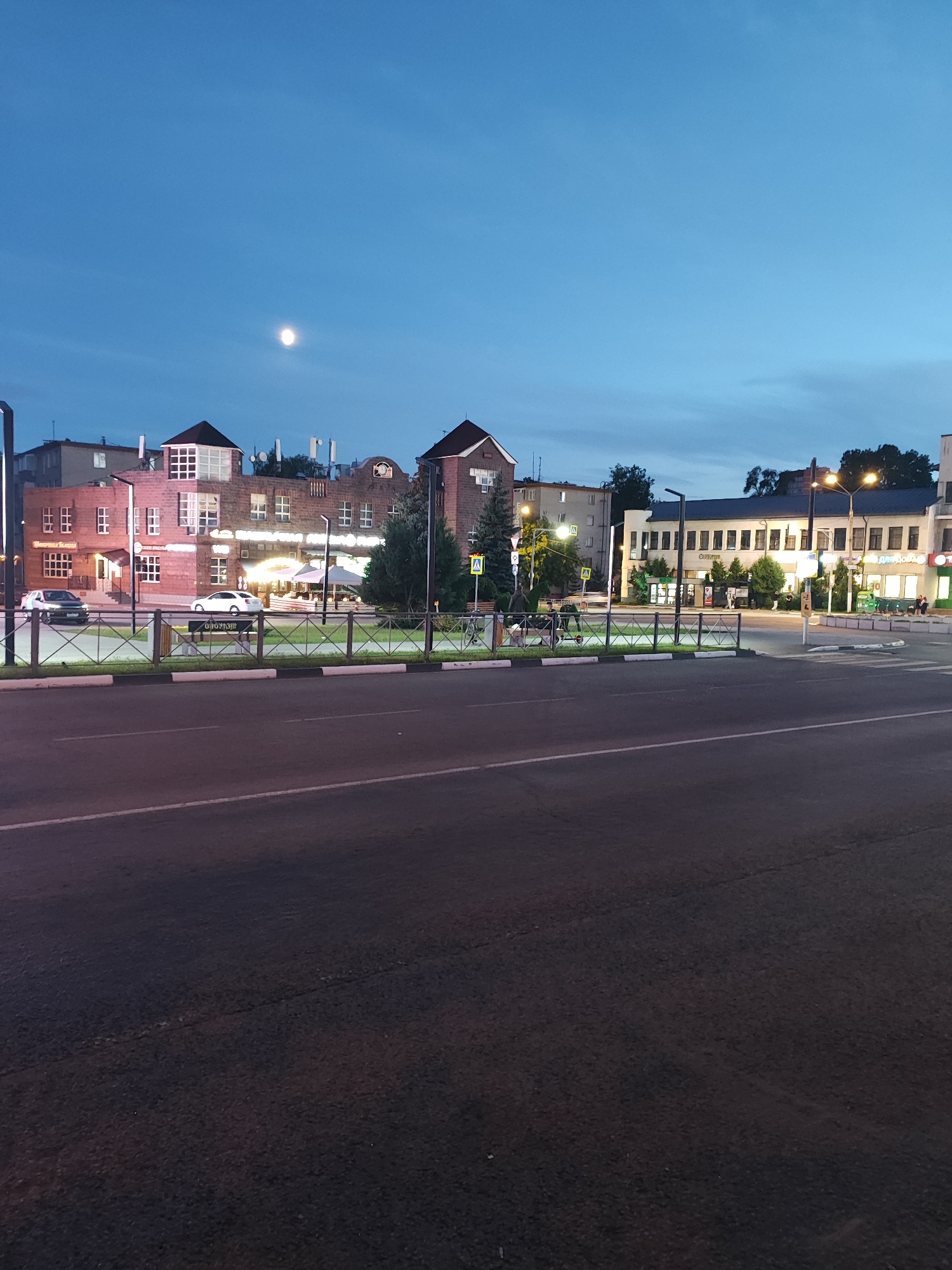 This media file depicts a twilight street scene in Shchyolkovo, Russia, with a clear, pale blue sky and a bright moon visible. The foreground is dominated by a dark asphalt road with white lane markings.
Behind the road, a pedestrian area is defined by a metal fence with a white top and black base, separating it from the street. This area features a paved walkway, a grassy strip, and a small hedge. Various objects are present, including bicycles, streetlights, and small trees.
The middle ground showcases a row of buildings. On the left, a brick building with multiple windows and a stepped roofline is visible, appearing to house businesses or residences. A white car is parked in front of it, and a dark SUV is driving past. Further to the right, a long, light-colored building with a dark roof is illuminated from within, suggesting it's open for business. Several signs are affixed to this building, though the text is largely illegible. The overall atmosphere is calm and serene, characteristic of early evening. The time of day is clearly indicated by the twilight sky and the artificial lighting of the buildings.
