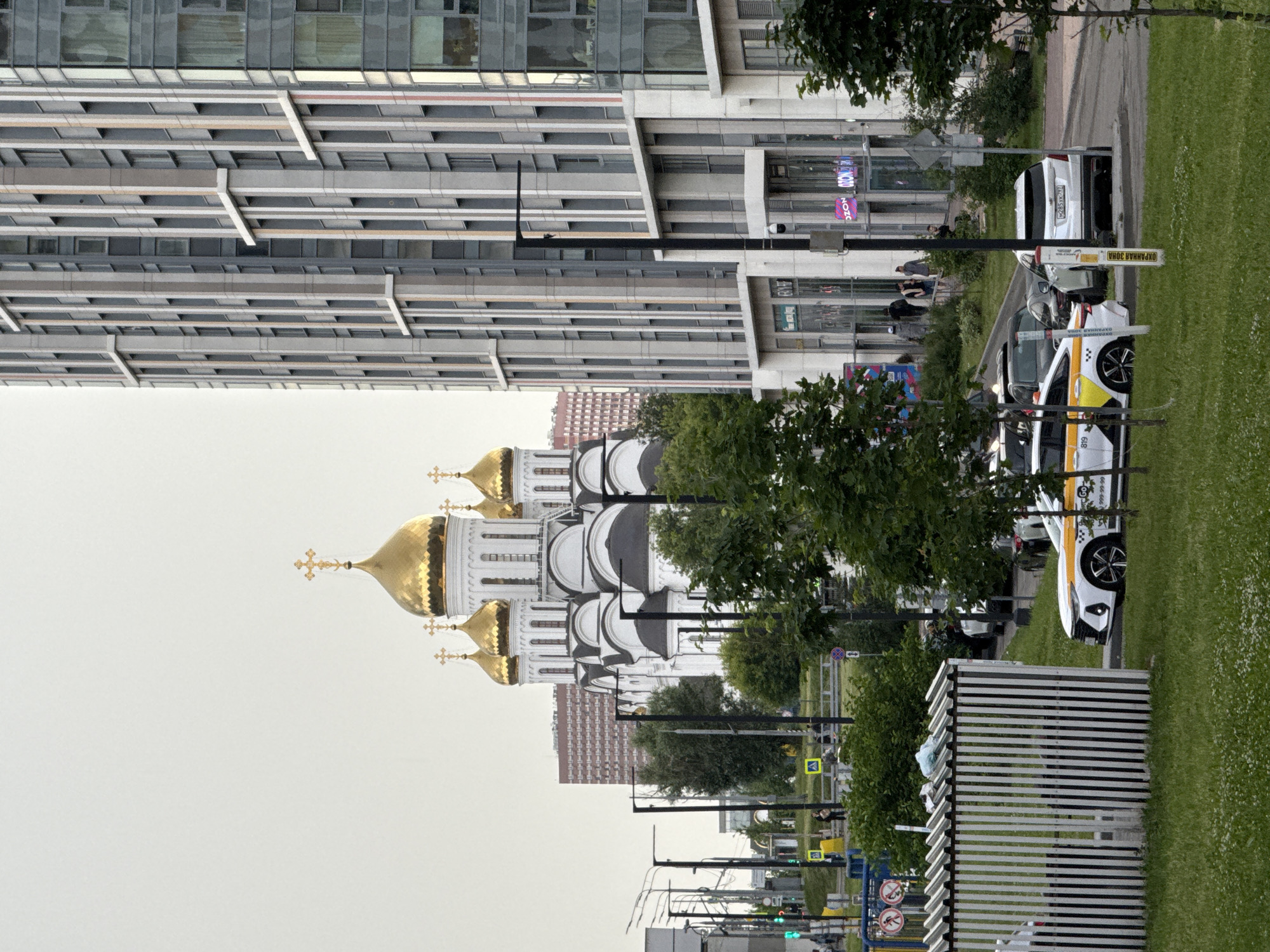 The image depicts a street scene in Moscow, Russia, featuring a modern high-rise building adjacent to a white church with golden domes. In the foreground, a white taxi with yellow and black markings is parked on the right side of the road. Several other vehicles are visible on the street, including a white SUV and a dark-colored car. A few pedestrians are present on the sidewalk in front of the building. The environment appears to be a city setting with trees and greenery along the roadside. The sky is overcast. The high-rise building has a contemporary architectural design with many windows. The church is characterized by its traditional onion domes, a common feature of Russian Orthodox architecture. The taxi has a phone number displayed on its side.