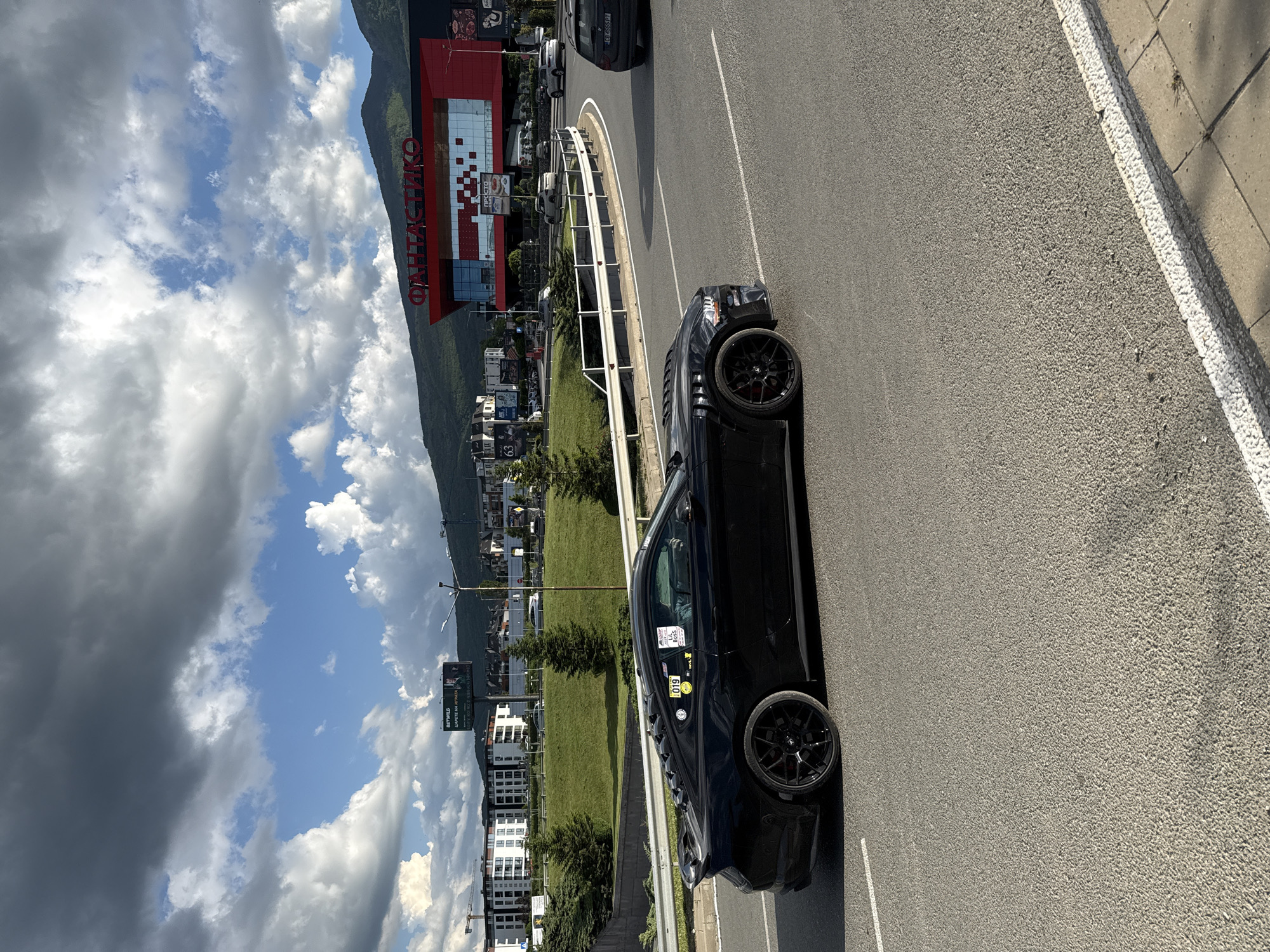 The image shows a black car parked on the side of a road in Sofia, Bulgaria. The location is identifiable by the large "ФАНТАСТИКО" supermarket building in the background and a mountain. The car is facing away from the camera. A sidewalk borders the road. The sky is partly cloudy.