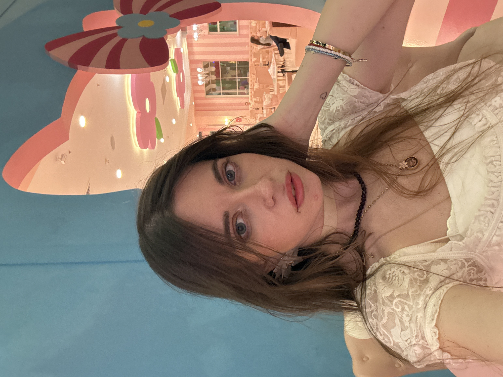 A young woman is taking a selfie in front of a backdrop decorated in a cartoonish style with pastel colors, reminiscent of a children's theme park or themed restaurant. The environment consists of pink and white striped walls, cut-out flowers, and a large, whimsical bow-shaped structure overhead. In the background, there is a glimpse of a brightly lit interior, possibly of a restaurant or event space with white furniture.
The woman has brown hair and is wearing a white lace top and multiple necklaces. Her arm is raised. The wall to the left is a solid light blue color. The photo was taken in the Hello Kitty-themed restaurant in Moscow, Russia.