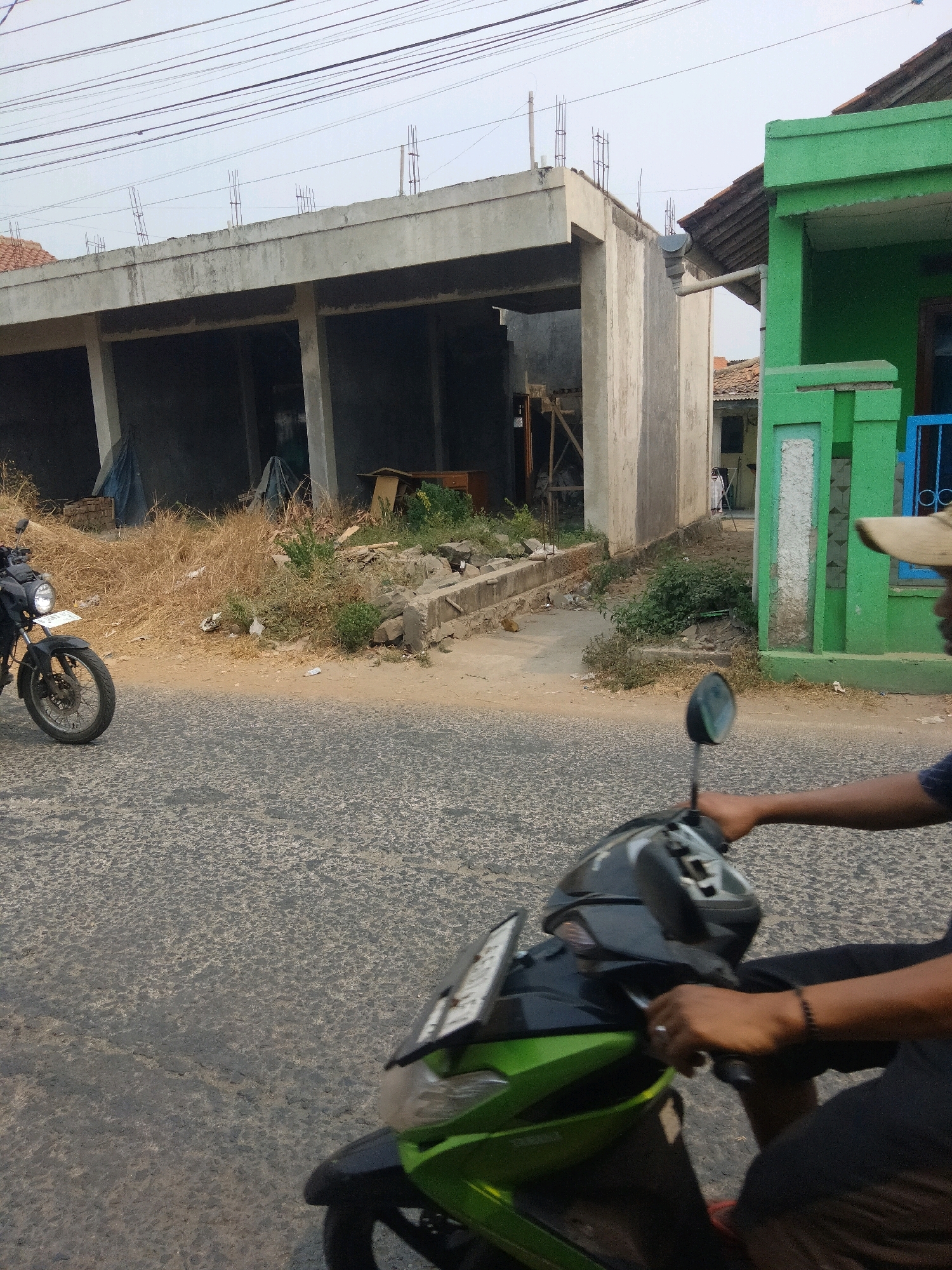 In the city of Suka Sari, Indonesia, a scene unfolds on a paved road next to a partially constructed building and a vibrant green house. The construction site features concrete pillars and beams, with scaffolding visible within its open structure. Dry grass and debris litter the ground near the building. A black motorcycle is parked on the left side of the road, its front wheel and handlebars in view. In the foreground, a person on a green and black motorcycle is captured from the chest down, with their hands on the handlebars. They are wearing black clothing and a tan baseball cap. The sky is overcast.