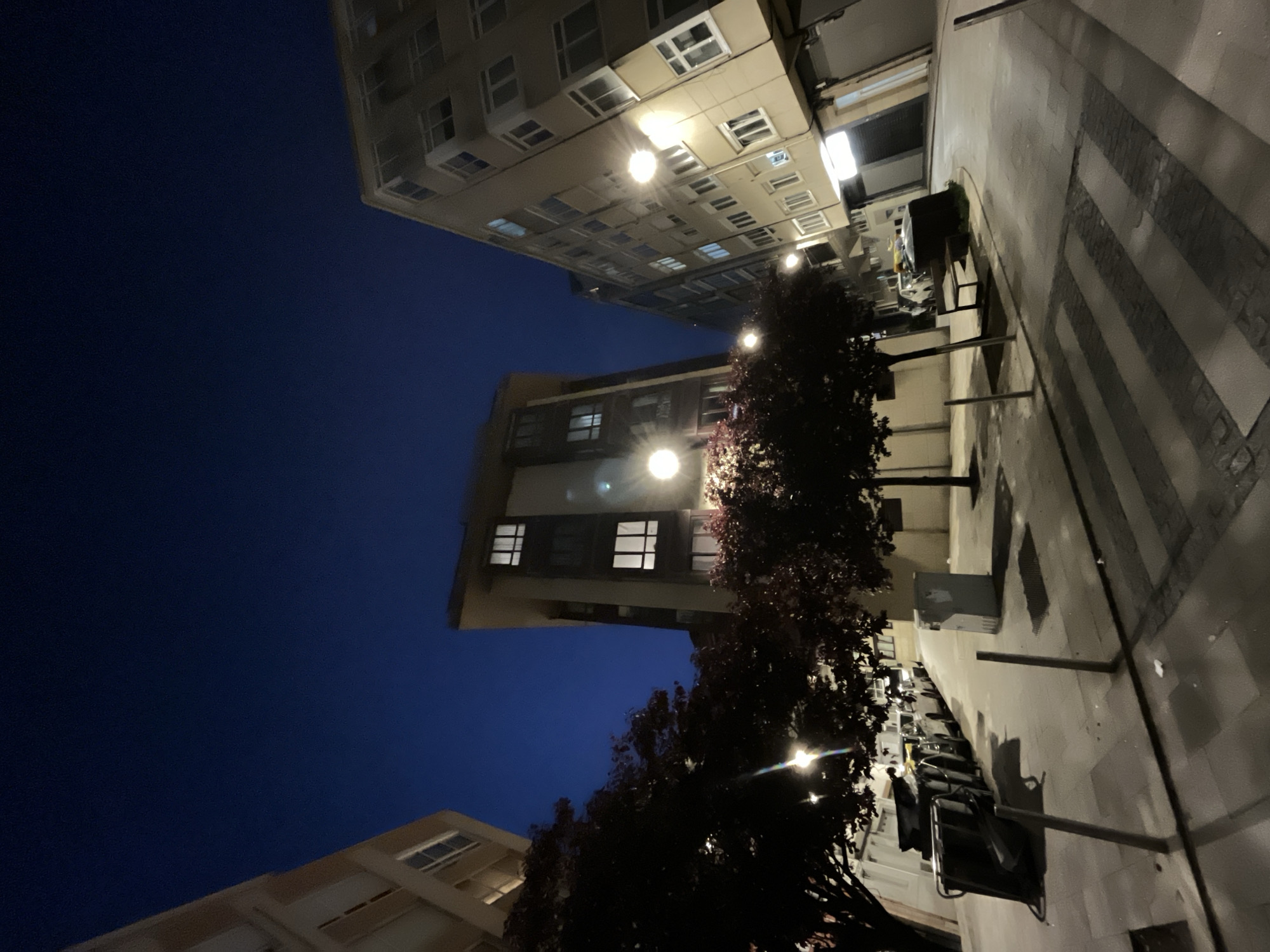 The image shows a street scene at night in A Coruña, Spain. Several tall buildings flank the street. The sky is a deep blue. Streetlights illuminate the buildings and the sidewalk, which is paved with light-colored stone and marked with darker stripes. Trees with dark foliage line the street. Benches and trash cans are visible along the sidewalk. The scene appears deserted, with no people visible.