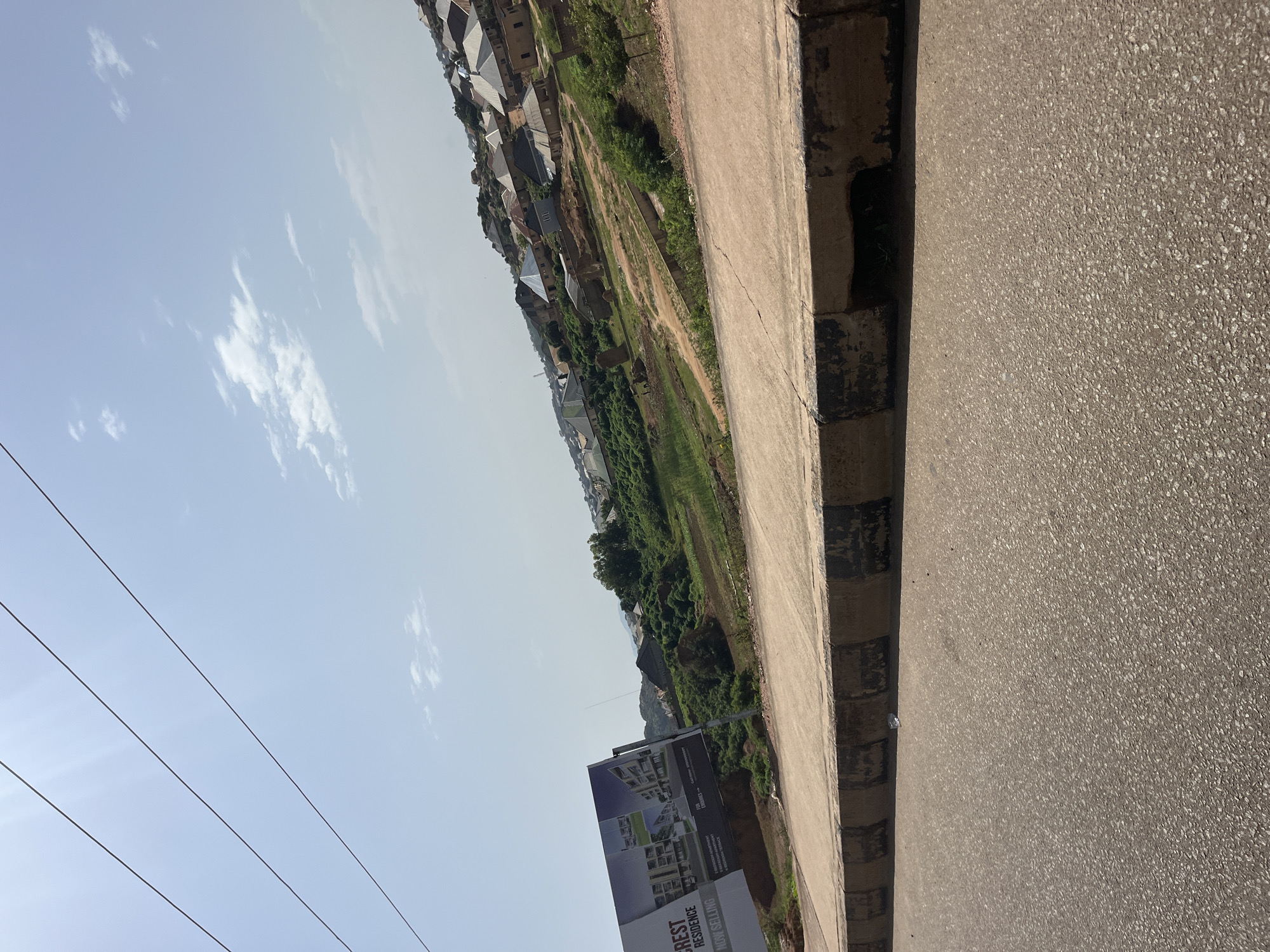 The image depicts a view of a residential area on a hillside in Bukuru, Nigeria. In the foreground, a paved road runs parallel to a concrete curb with black and beige markings. A large billboard advertising "CREST RESIDENCE" is tilted and angled to the left, displaying images of modern apartment buildings and the text "NOW SELLING". Beyond the road and billboard, the hillside is covered with a mix of green vegetation and scattered residential buildings with corrugated metal roofs, suggesting a developing urban or suburban environment. The sky above is clear with a few scattered clouds, and several power lines are visible against the light blue sky. The overall scene suggests an overview of a community under development, with an emphasis on residential construction and infrastructure.