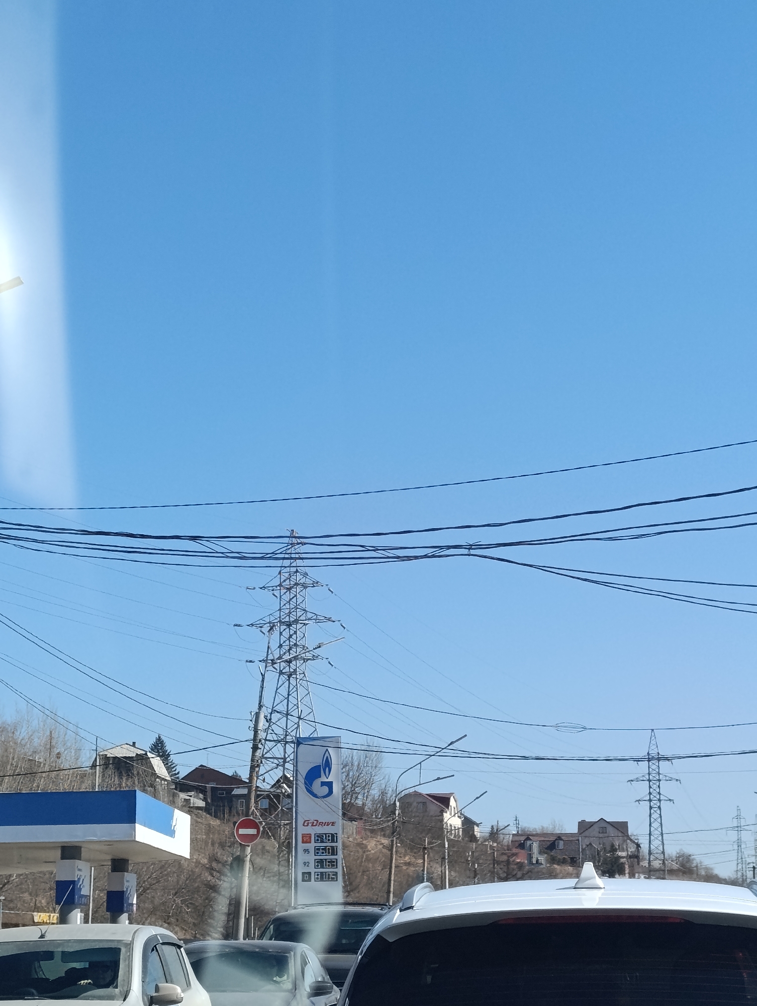 The image shows a street scene in Krasnoyarsk, Russia, with a Gazprom gas station in the foreground. The gas station's sign displays prices for 95, 92, and diesel fuel. Several cars are visible in the foreground, including a white car. Overhead, power lines crisscross the sky. In the background, there are residential buildings on a hill. A red "No Entry" traffic sign is located near the gas station. A transmission tower is visible near the gas station and more towers are in the distance. The sky is clear and blue.