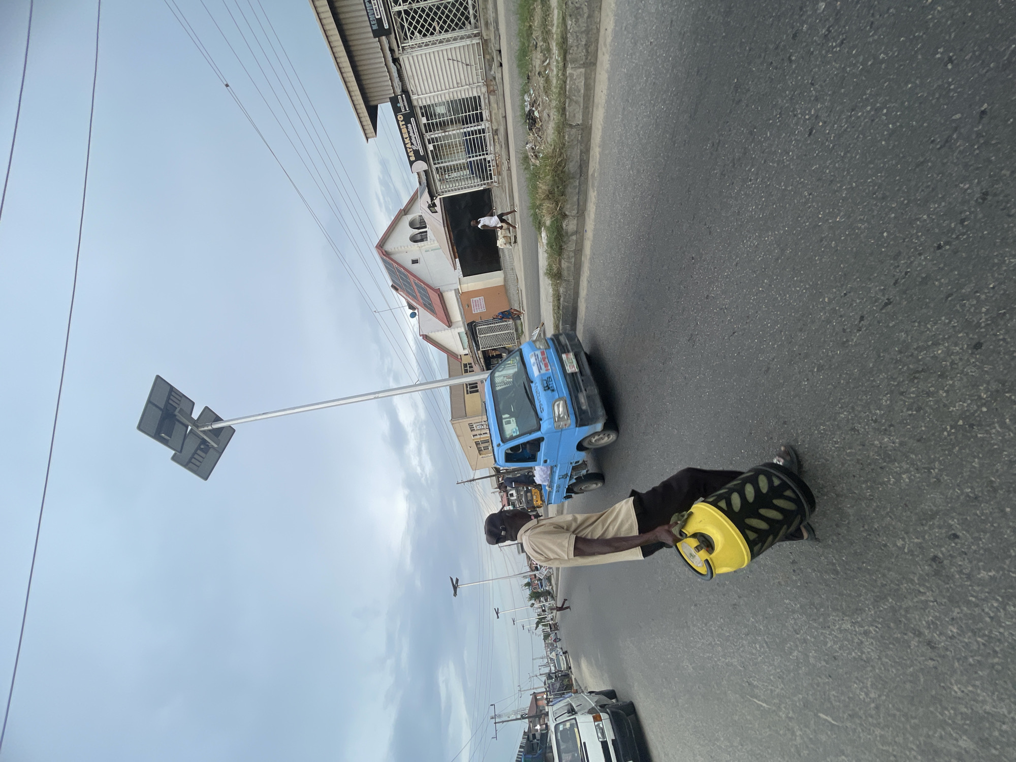 The image depicts an urban street scene in Ado, Nigeria, under an overcast sky during daytime. A man is the central subject, walking on a paved asphalt road. He wears a light-colored short-sleeved shirt, dark pants, and a dark cap. He is carrying a distinctive yellow and black gas cylinder, which has a yellow top and base and a black, possibly caged or textured, body.
In the mid-ground, a light blue mini-truck or utility vehicle is visible on the street, behind the man. Further down the street, a white vehicle is partially seen on the left, and other vehicles and people are indistinctly visible in the distance. Utility poles and wires crisscross the sky. A prominent street light or signpost with a square, dark panel is on a tall pole to the left of the main subject, and similar poles extend down the street.
Along the right side of the street, several commercial-looking buildings line the road. One notable building has a white facade, a gabled red roof with what appear to be solar panels, and arched windows. Another building features a large roller shutter door and signs. Text visible on one building includes "KEMPYA" and "SAYAKETTO". Another person in a white shirt and dark shorts is seen walking near the buildings. The roadside verge consists of dirt, sparse grass, and some debris. The scene captures the daily activity and commerce of a street.