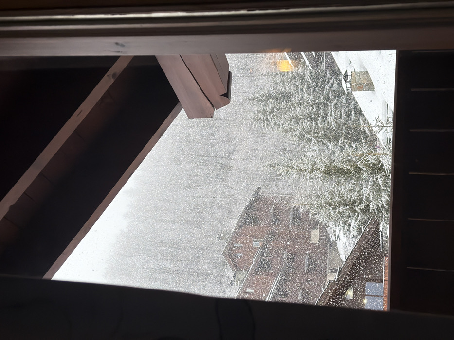 The image displays a winter mountain landscape in Les Allues, France, viewed from a room or balcony during active snowfall. In the immediate foreground, a dark wooden balcony railing is visible along the bottom edge, with a dark wooden overhead structure, including exposed beams, framing the top and right sides of the view. Heavy, visible snowflakes are falling across the entire scene.
In the midground, a prominent multi-story building with a dark reddish-brown exterior, multiple windows, and a steep, snow-covered roof is situated on the left, partially obscured by the falling snow. Below and to its right, the snow-covered roof of another building is partially visible. Numerous evergreen trees, heavily blanketed in snow, are interspersed throughout the midground and extend up the hillside. A stone chimney with a conical metal cap emerges from the snow-covered ground on the right side of the frame. The background consists of a densely forested mountain, completely covered in snow, appearing hazy due to the ongoing snowfall. No individuals or animals are present in the scene.