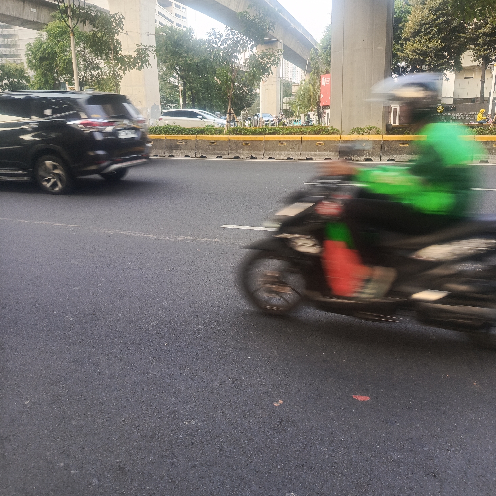The image captures a motion-blurred view of a busy street in South Jakarta, Indonesia, under an elevated overpass. A black SUV is visible on the left, its rear end blurred due to motion. In the foreground, a motorcycle is moving from right to left, also heavily blurred. The rider is wearing a bright green jacket and a helmet, suggesting they might be a delivery driver or courier. The street is paved with asphalt, marked by a white line. Concrete barriers with yellow markings separate the roadway from a planted median strip with trees. In the background, buildings and more trees are visible. The overall impression is one of urban movement and activity.