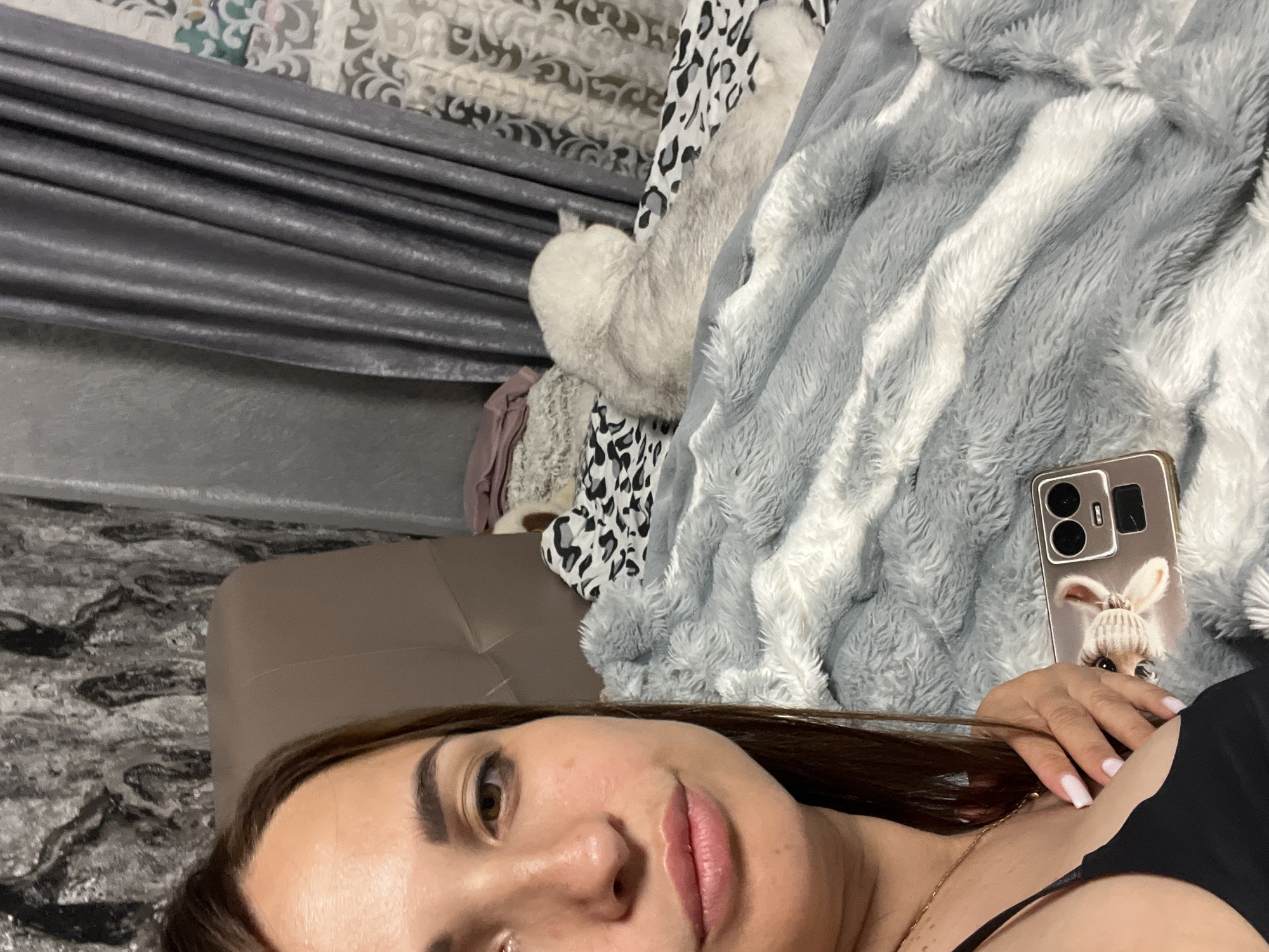 A woman is lying on a bed, looking at the camera. She has brown hair and is wearing a black top. On the bed, there is a fluffy gray and white blanket, a phone with a picture of a dog on the case, and a white dog lying down. The room has gray curtains and a patterned wallpaper.