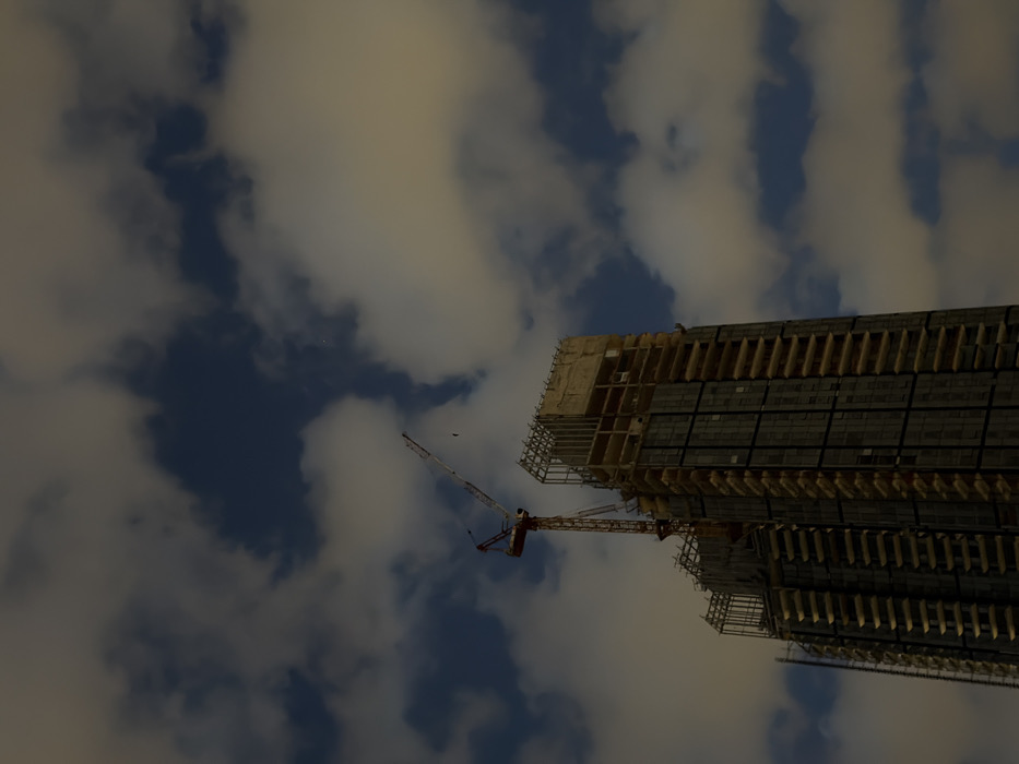 A tall building under construction reaches toward a cloudy sky. The building's skeletal frame is visible, punctuated by numerous rectangular openings. A red and white crane is attached to the building, likely aiding in the construction process. The clouds fill the majority of the frame, creating a soft, atmospheric backdrop. The angle of the shot looks upwards, emphasizing the height of the building.