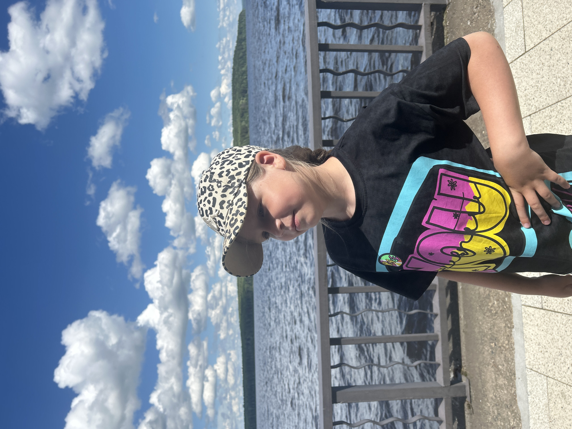 A young person wearing a black t-shirt with a colorful graphic and a leopard-print baseball cap stands outdoors, leaning against a railing. The person's head is tilted down, and they are looking towards their right. The t-shirt graphic is prominent, featuring large, stylized letters in yellow, pink, and purple, with smaller colorful graphics within the letters.
The scene is set near a large body of water, likely a river or lake, with ripples on the surface indicating a slight breeze. In the background, a tree-lined shore is visible, suggesting a natural landscape. The sky is bright blue with scattered white, fluffy clouds, indicating a clear and sunny day. The lighting suggests it is daytime, possibly late morning or early afternoon.
A metal railing runs along the edge of the paved area where the person is standing, separating it from the water. The ground appears to be a light-colored paved surface. The overall mood seems relaxed and casual. No specific event is indicated, other than enjoying an outdoor setting by the water. The location is identified as Chaikovsky, Russia.
