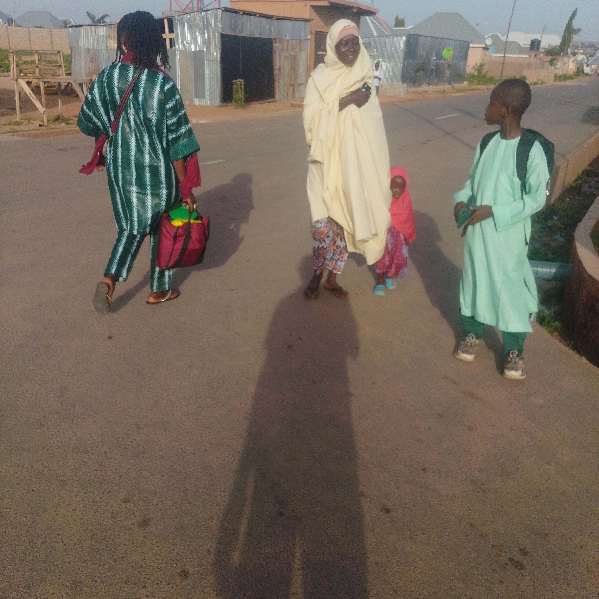 In an outdoor urban environment, likely a street, two adults and two children are walking. On the left, a person wearing a green and white striped garment with a red scarf and a maroon bag is walking away from the camera. To their right, a woman in a cream-colored hijab and patterned pants is walking with a young child in a pink outfit and blue shoes. On the far right, a young boy in a mint green tunic and dark pants, carrying a backpack, is walking towards the camera. The scene is set against a backdrop of buildings with corrugated metal and tiled roofs, suggesting a developing or informal urban area. The ground is paved, and shadows indicate it is daytime, with the sun casting long shadows across the street.