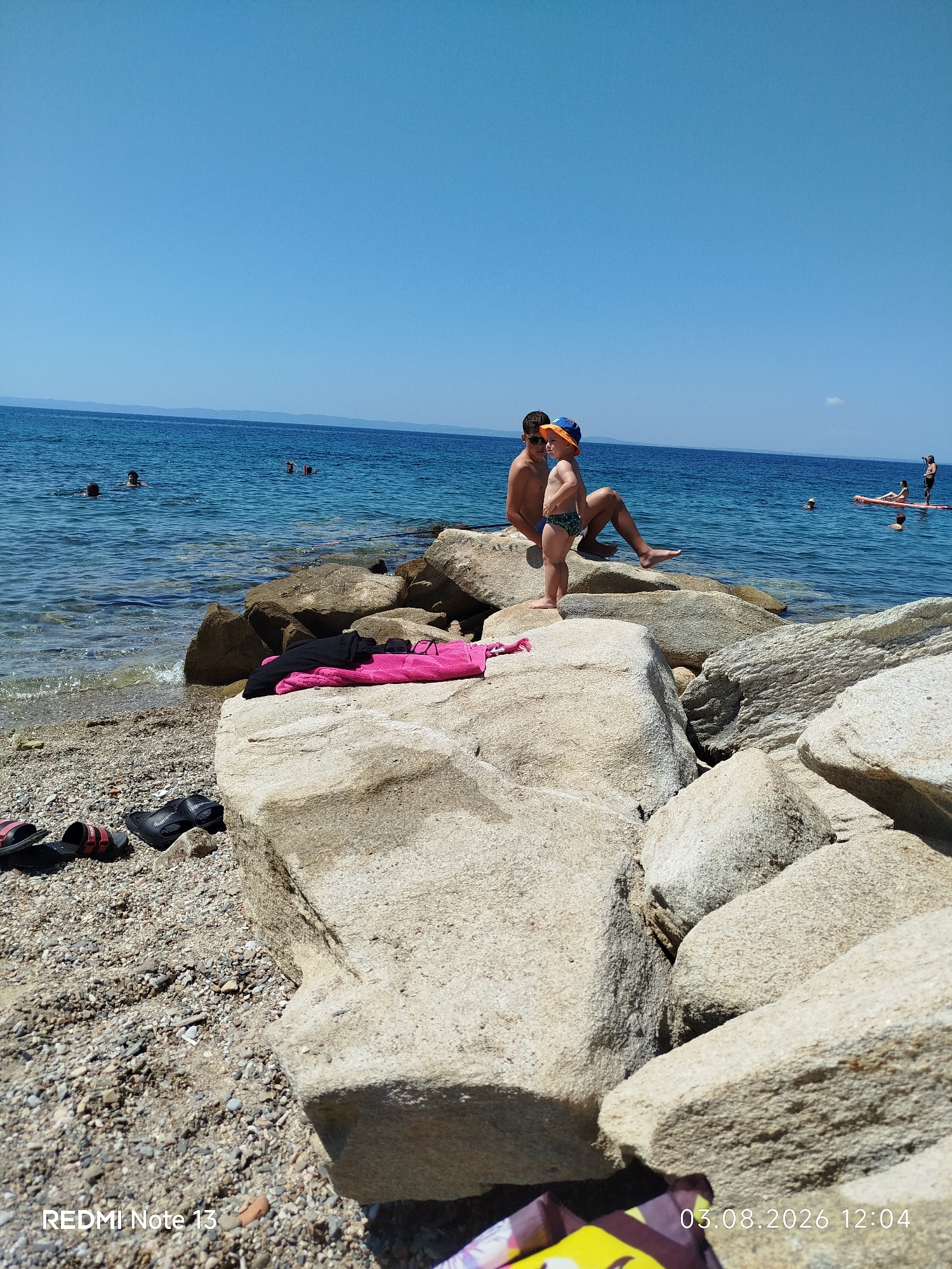 A high-angle, long shot shows a rocky beach scene with clear blue water and a bright blue sky. In the foreground, large, light-colored rocks are scattered on the shore. A pink towel and black clothing are placed on one of the rocks. Several pairs of flip-flops are visible on the pebbly ground to the left.
Further back, two young boys are seated on a large rock near the water's edge. The older boy, with dark hair, is looking away, while the younger boy, wearing an orange hat and patterned swim trunks, is looking towards the older boy.
In the water, several people are visible, some swimming and others on a paddleboard in the distance. The water is calm and translucent near the shore, revealing the rocky seabed. The scene appears to be a sunny day at a Greek beach.