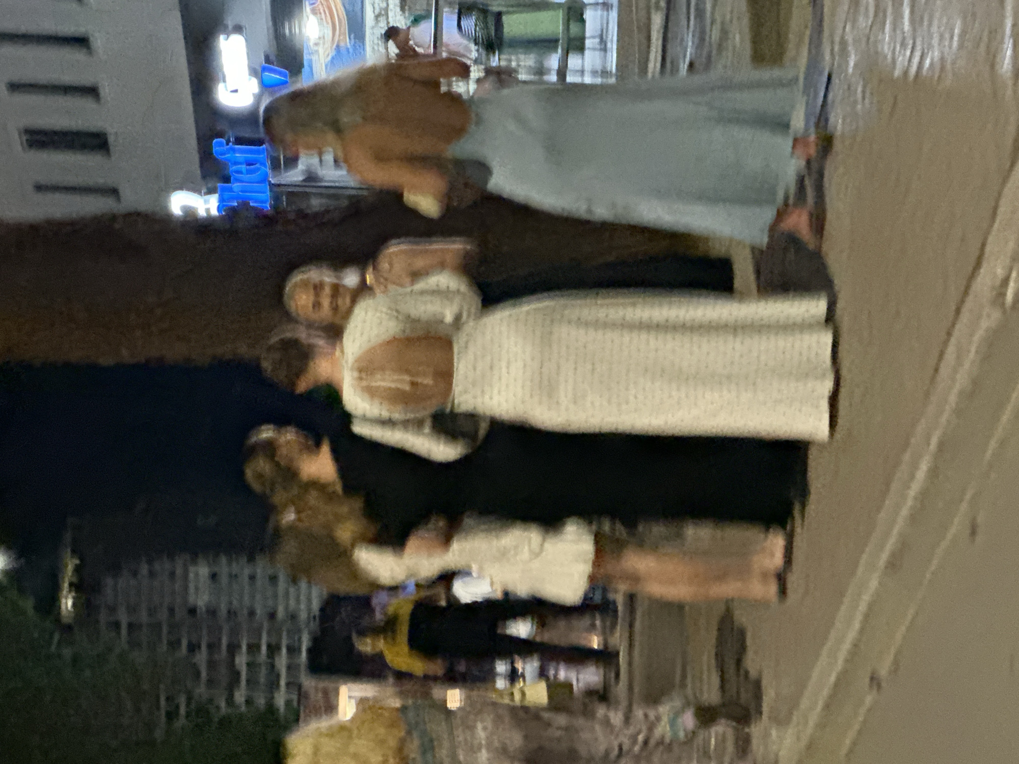 In Budva, Montenegro, a group of five women, dressed in formal attire, are standing on a paved area outside at night. The woman in the foreground on the right wears a light blue, backless evening gown. To her left, a woman in a white, polka-dotted backless gown stands with her back to the camera, talking to a woman in a black dress and a woman in a white dress. Further left, another woman in a patterned dress is partially visible. In the background, a man in a yellow and black shirt walks away from the group. The scene appears to be near a building with illuminated signage that reads "chef" in blue and white. The environment is a street at night, with a dimly lit brick building visible in the upper left corner.
