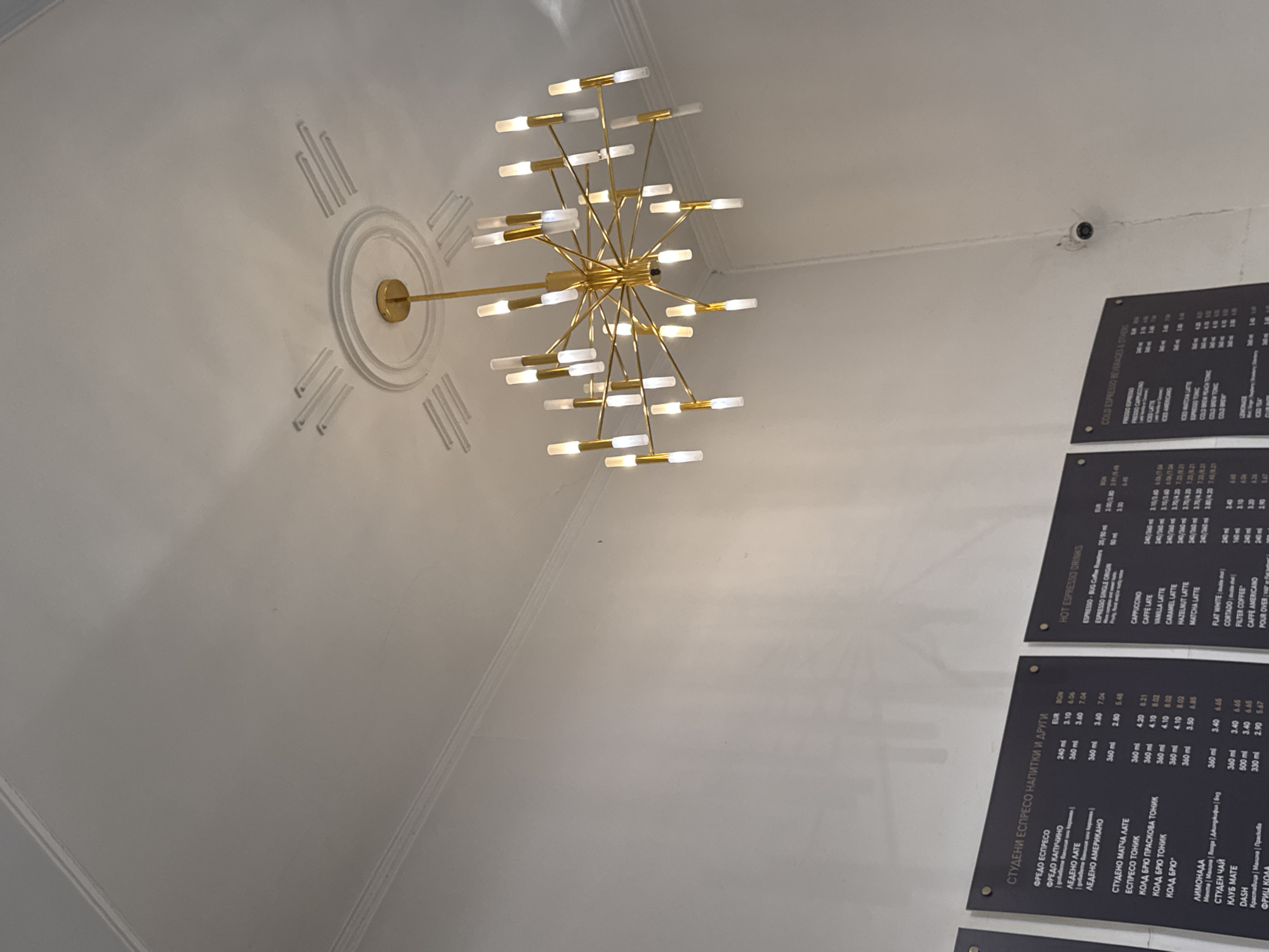 The image shows the interior of a cafe, likely in Sofia, Bulgaria, given the Cyrillic text on the menus. The primary focus is a modern, multi-tiered brass chandelier with numerous glass tube light fixtures, hanging from a white ceiling with ornate decorative molding. Two black menu boards are visible on the left and right sides of the frame, displaying drink options in Cyrillic and Latin script, respectively. The menu boards list various espresso drinks, lattes, and other beverages with sizes and prices in EUR and BGN. The walls are painted white, and a white security camera is mounted high on the wall on the right side of the image. The overall lighting is soft, primarily from the chandelier, creating a warm ambiance. There are no people visible in the frame.