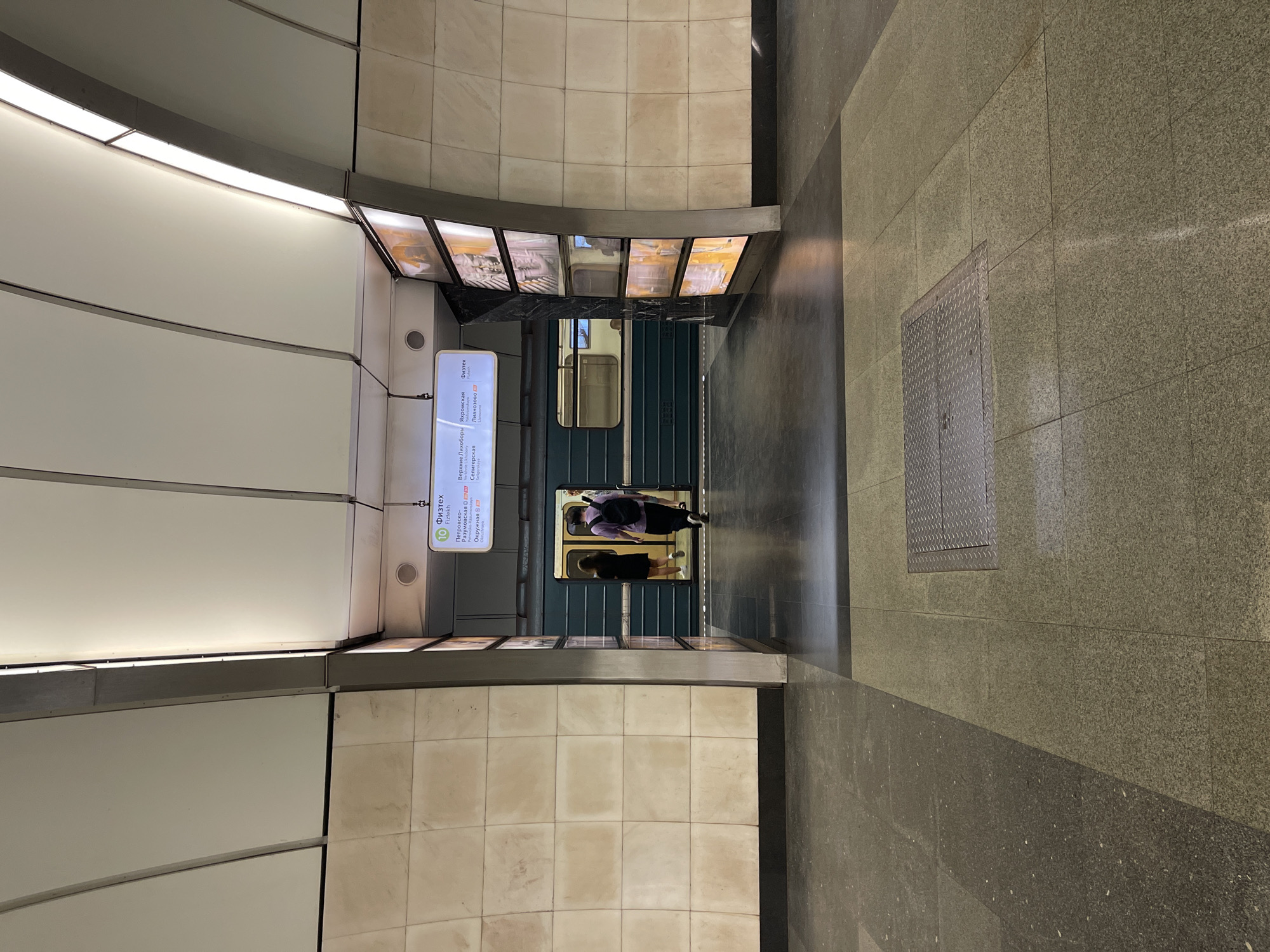 The image depicts an interior scene of a metro station. The perspective is from above, looking down onto the platform and train.
The station features curved walls tiled with a light cream color. Above the platform edge, there is a display with station names in Russian. A train is visible at the platform, its doors open. Two people are visible standing near the train doors. The platform floor is tiled with a combination of light and dark gray tiles. There's a metallic grate on the floor. The scene is well-lit with overhead lighting.
The location is a metro station in an unknown city.
