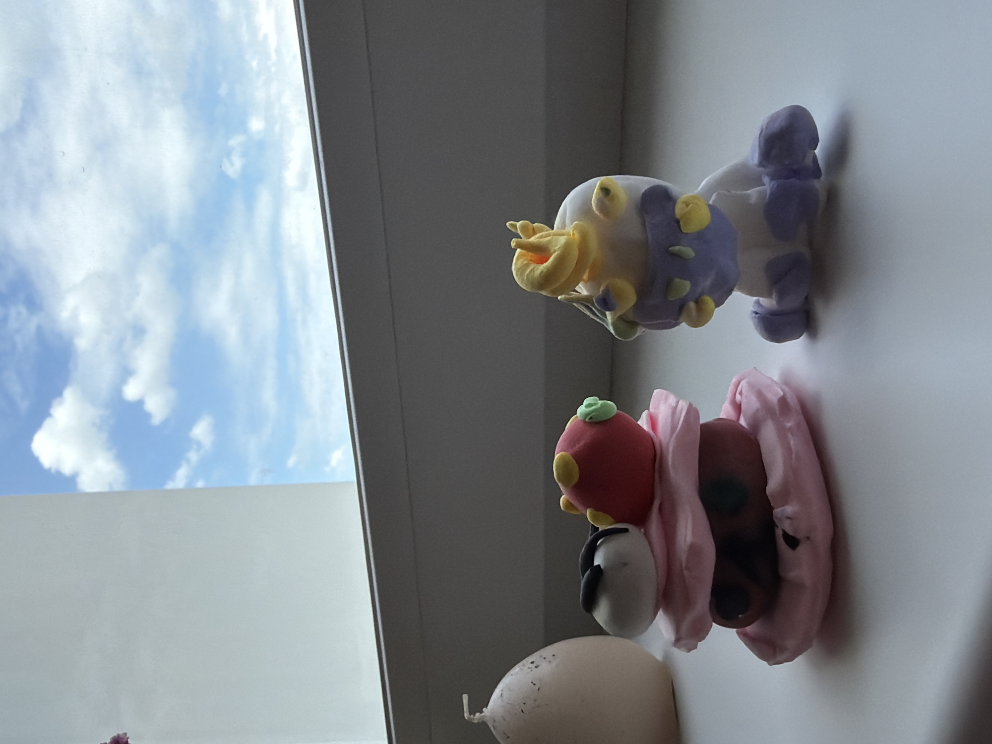 An outdoor shot shows a windowsill with two clay figures and a candle. The candle, on the left, is tall and oval-shaped with a cream color and a dark speckling. The first clay figure, a stack of pink and brown circular pieces, is on the right of the candle. The second clay figure is on the right of the stacked figure; it is a squat, white figure with purple accents, including a purple band around its head, and yellow accents on its head and shoulders. The windowsill is light gray and is set against a bright blue sky with fluffy white clouds.