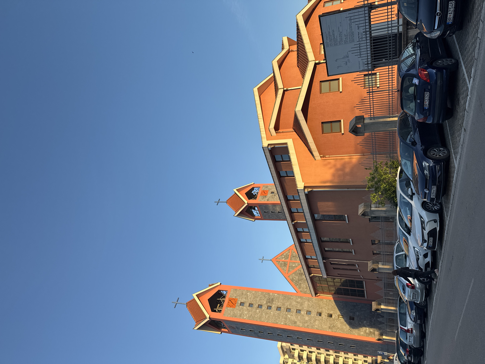 The image, oriented 90 degrees counter-clockwise from its original capture, displays a modern cathedral in Bar, Montenegro, and a line of parked cars under a bright, clear blue sky. The main building, identified as a cathedral, features a distinctive reddish-orange facade with tiered rooflines and grey stone accents. It has two prominent bell towers, each topped with a cross and equipped with a clock face. Numerous rectangular windows are visible across its walls.
A row of cars is parked along the right side of the frame. Several Montenegrin license plates are discernible, including "BR AZ546," "BR CE528," "BR YYK 759," "BR DM060," and "BR CM 094," where "BR" denotes Bar. A person is visible standing near a silver parked car.
In front of the main building, behind a fence, stands a large information sign. Visible text on this sign clearly states "KATEDRALA SV. PETRA – BAR," confirming it as the Cathedral of St. Peter in Bar. The scene is illuminated by bright sunlight, casting long shadows from the buildings and vehicles, suggesting either morning or late afternoon. The sky is entirely cloudless, indicating fair weather. The photograph captures an urban environment centered around a significant religious edifice.