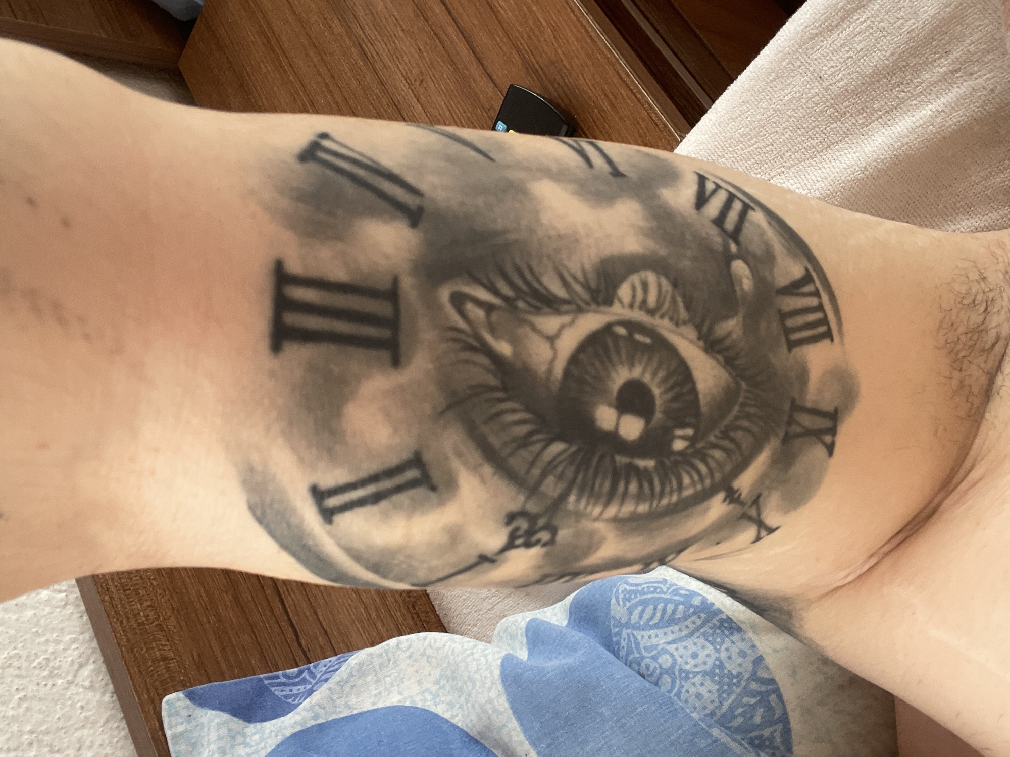 A close-up shot captures a person's light-skinned arm or leg, prominently featuring a large, intricate tattoo. The tattoo depicts a realistic human eye at its center, surrounded by a clock face with Roman numerals. The eye has a detailed iris and visible eyelashes, with a small reflection or highlight in the pupil. The clock face displays Roman numerals from I to XII, though not all are fully visible due to the eye's placement and the limb's curvature. Visible numerals include I, II, III, IV, VII, VIII, IX, X, XI, XII. The tattoo style is black and grey, with soft shading suggesting clouds or smoke around the clock face, giving it a somewhat ethereal or dreamy quality.
The limb rests on or near various household items. To the left, a section of a wooden surface with a distinct grain pattern is visible, likely part of furniture like a table or headboard. On the opposite side, a textured, off-white or cream-colored fabric, possibly a towel or blanket, is partially seen. Below the tattooed limb, a blue and white patterned fabric, perhaps another blanket or piece of clothing, adds a touch of color. A small, black electronic device, possibly a remote control or a charger, is subtly visible nestled between the wooden surface and the tattooed limb. Some hair is visible on the skin to the right of the tattoo, and a few small moles or freckles are visible on other areas of the limb. The lighting suggests an indoor setting, and the focus is entirely on the limb and its elaborate tattoo.