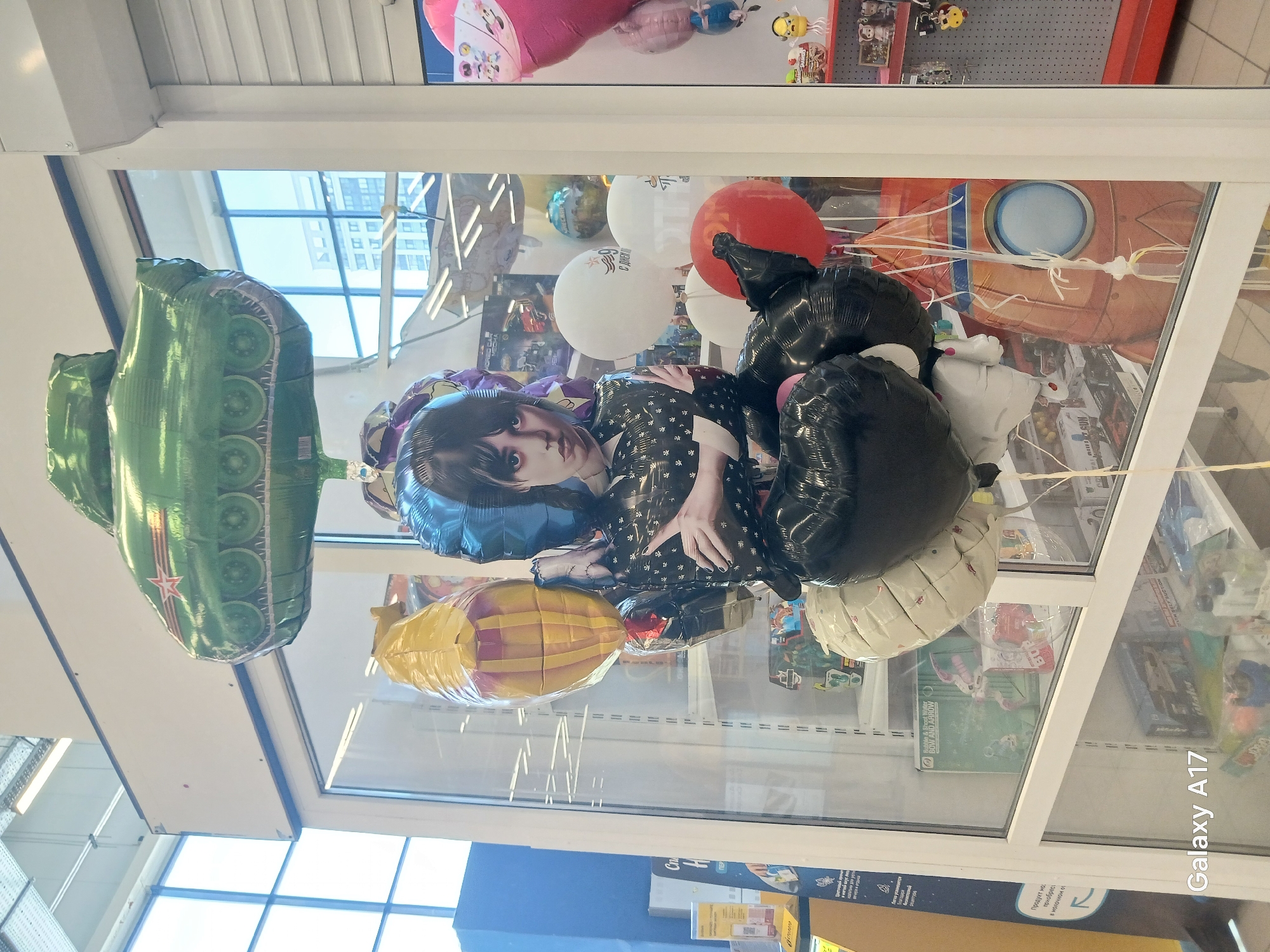 The image depicts a collection of inflated foil balloons hanging from the ceiling of a retail store. The balloons are various shapes and designs, including a green tank, a yellow rocket, balloons in the shape of the character Wednesday Addams, as well as black, red, and white balloons. The balloons are suspended by strings that are attached to the ceiling.
The photograph was taken inside the store, as can be deduced by observing a window that reflects elements of the city of Yekaterinburg, Russia. Visible outside the window are buildings. Inside the store, the reflection of the balloons and store merchandise on display, like board games, can be seen in the window. A sign is visible, printed in Russian, which suggests the location is Russia. At the bottom right of the image, "Galaxy A17" is superimposed.