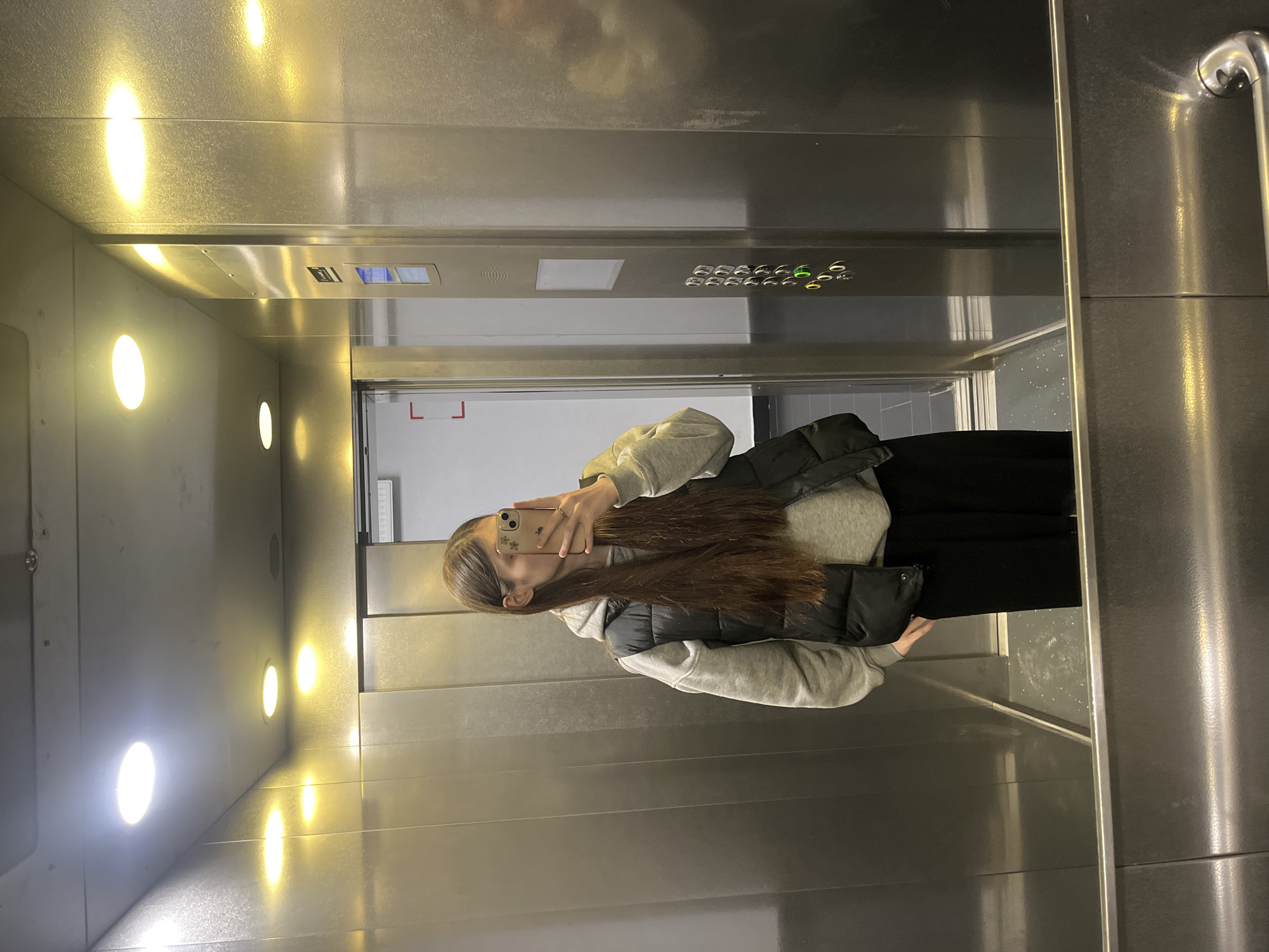 A young woman with long, brown hair is taking a mirror selfie in an elevator. She is wearing a light gray hoodie and a dark-colored vest or jacket. The elevator has stainless steel walls with illuminated buttons and round lights. The floor indicator lights are visible above the door. The buttons indicate various floor levels, with the '6' button illuminated. The elevator is located in Saint Petersburg, Russia. The environment is clean and modern. The woman is holding a smartphone. The elevator doors are partially open, revealing a glimpse of the building interior.