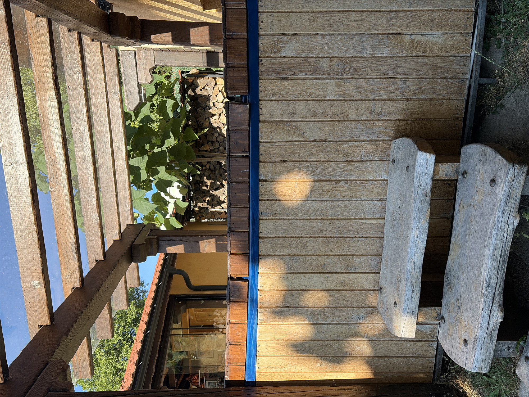The image depicts an outdoor seating area, likely part of a restaurant or a guesthouse in the city of Smolyan, Bulgaria. In the foreground, two rustic wooden benches made from rough-hewn logs are positioned in front of a tall wooden fence. The fence is constructed from vertical planks of light-colored wood. Above the fence, a bright blue band is visible. The background reveals a wooden pergola with a transparent roof, allowing sunlight to filter through. Beyond the pergola, a building with an orange-tiled roof and yellow walls can be seen, along with a large green tree. A stack of firewood is also visible in the background. The scene is bathed in sunlight, casting shadows on the wooden fence. There are no people present in the image.