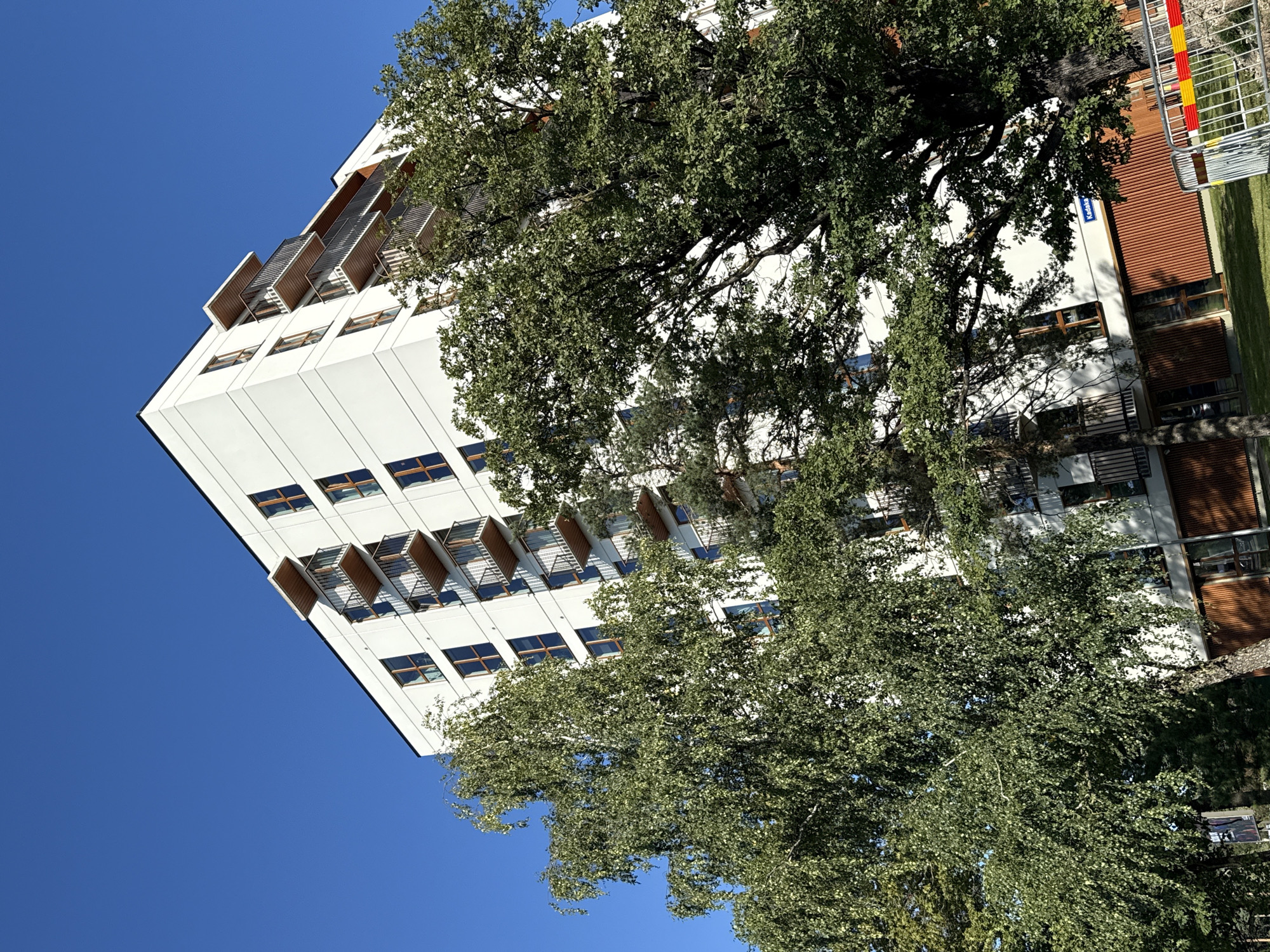 In Tallinn, Estonia, a modern white apartment building with multiple balconies and large windows stands tall against a clear blue sky. The building is partially obscured by lush green trees, one with delicate, hanging branches and another with a broader canopy, creating a contrast between the urban structure and the natural landscape. At the base of the building, a wooden facade is visible, and a section of a metal fence with red and yellow warning stripes suggests ongoing construction or a temporary barrier. A street sign indicating "Kadaka" is also visible on the building. The scene depicts a contemporary residential area integrated with greenery.