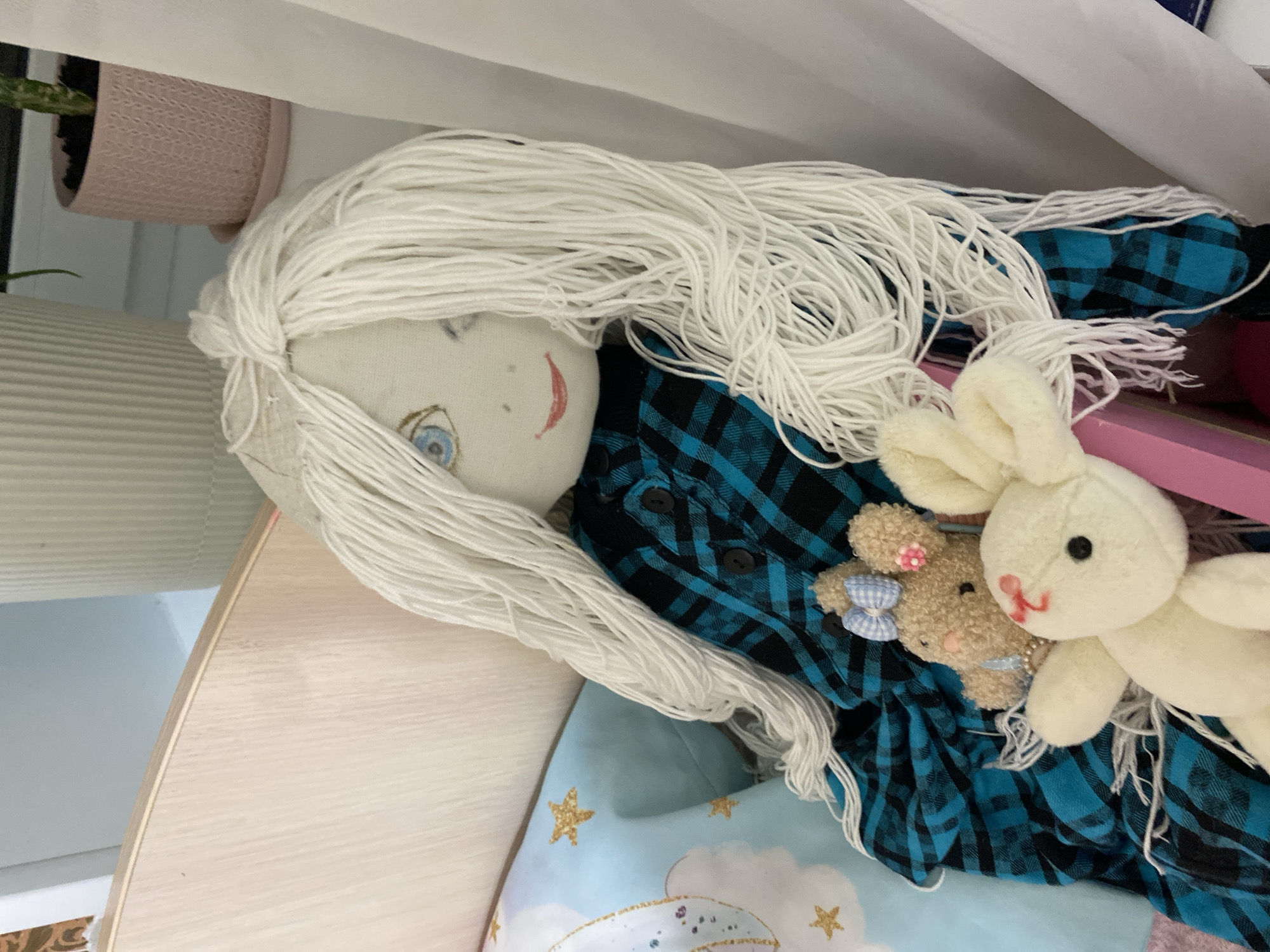 The image depicts a handmade doll with long, white yarn hair and painted facial features, including blue eyes and red lips. The doll is dressed in a blue and black plaid shirt. It is sitting upright, leaning against a light-colored wooden surface, possibly a bed frame. To the left of the doll is a light blue pillow with gold stars and clouds. In front of the doll, there are two stuffed animals: a small, fuzzy brown bunny with a blue bow and a pink flower, and a larger, cream-colored bunny with red markings on its face. The background shows a vertical ribbed object, a pink planter with a plant, and white fabric curtains. The scene appears to be indoors, likely in a bedroom. There is no specific indication or evidence of this scene being located in Lomonosov, Russia.