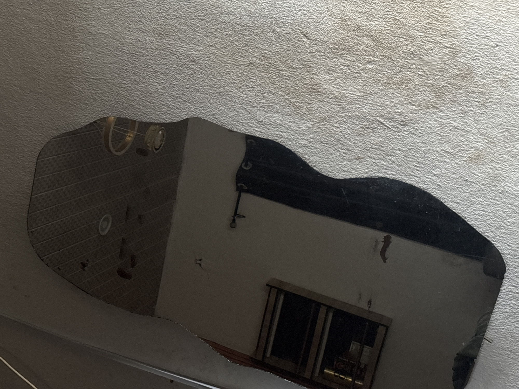 The image shows a portion of a mirror reflecting a room interior. The reflection includes a window with dark curtains, part of a wall, and possibly a section of ceiling with light fixtures or ventilation. The mirror is mounted on a textured surface, possibly a ceiling. The mirror's shape is irregular, suggesting it may be a custom or fragmented piece. The location in Ikot Ekpene, Nigeria cannot be determined from the provided image.