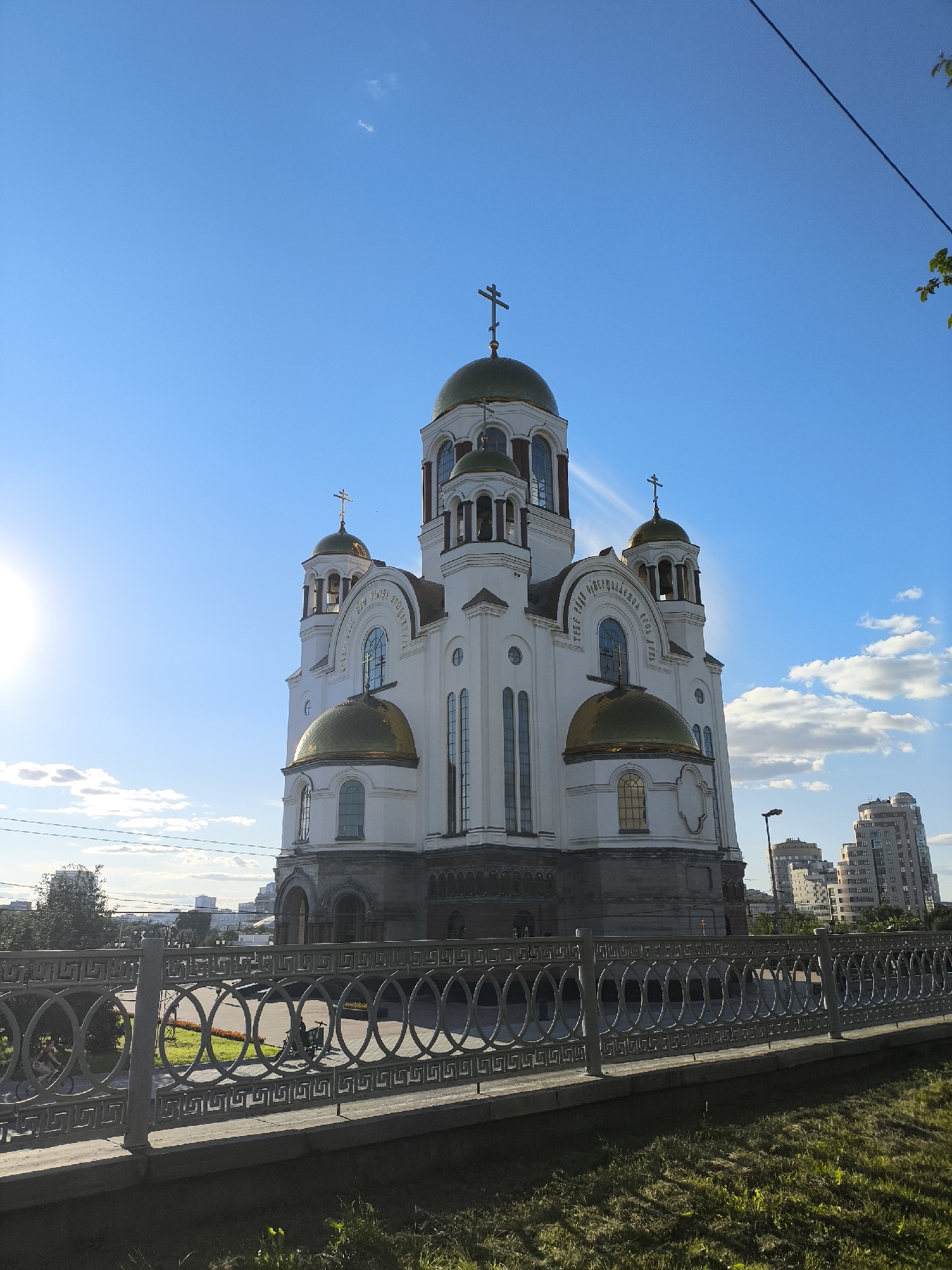 The image shows the Church on Blood in Yekaterinburg, Russia, on a clear day with a bright sun. The white church features golden domes and crosses, with arched windows and ornate architectural details. In the foreground, a decorative metal fence runs along a paved walkway. Behind the church, modern buildings and trees are visible, indicating an urban setting. The sky is a vibrant blue with a few scattered white clouds. A lens flare from the sun is prominent on the left side of the image. There are no discernible people or specific actions depicted in the frame.