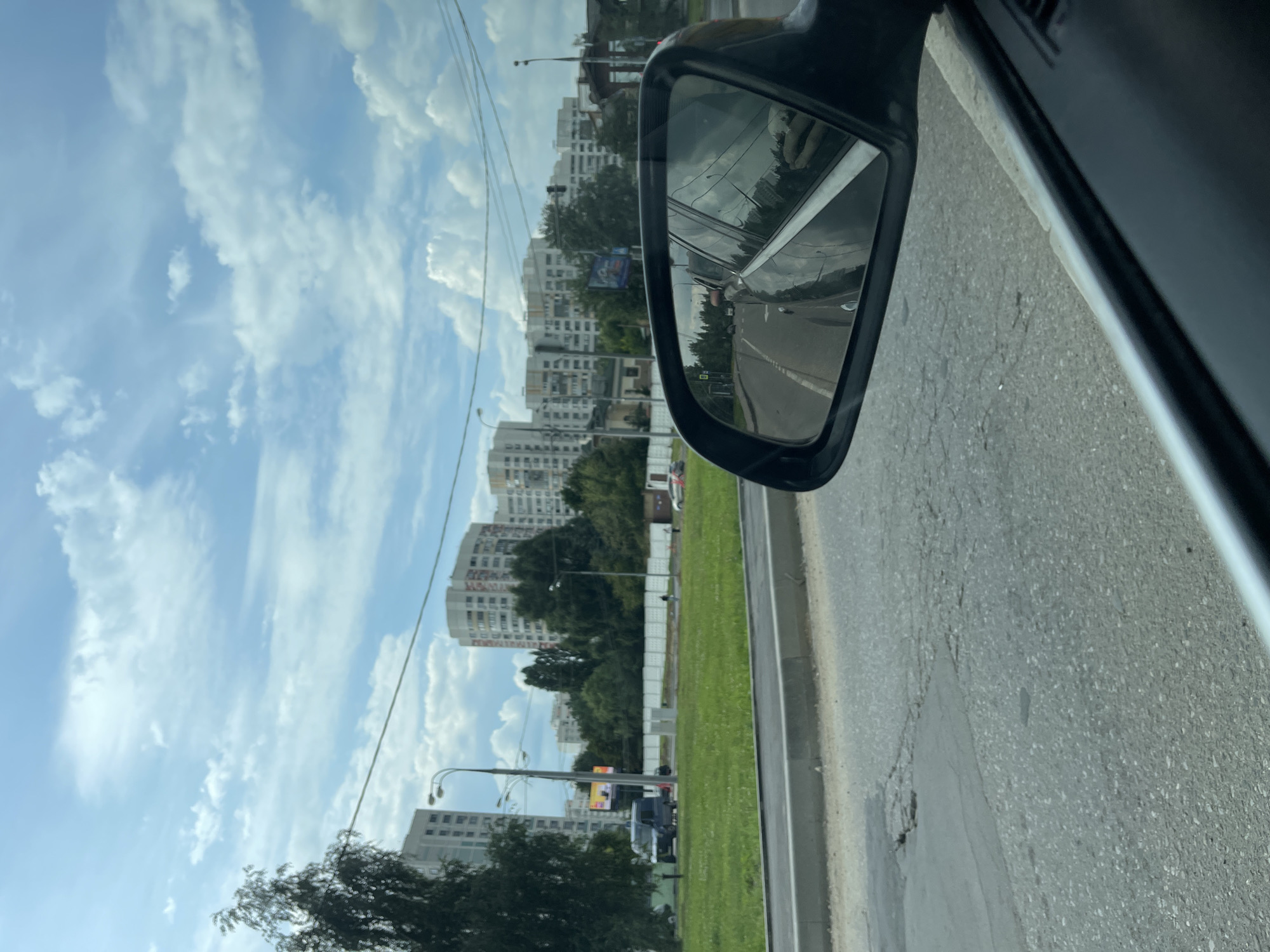 The image captures a view from inside a vehicle, looking forward and towards a side mirror, on a partly cloudy day in Moscow, Russia. The foreground shows a section of asphalt road, which appears worn with patches of repair. To the left of the road is a green grassy area bordering a sidewalk. Beyond the grass, a row of tall, white apartment buildings, characteristic of Moscow's urban landscape, stretches across the background. Trees are interspersed between the buildings and in front of them. A few vehicles, including a dark van and a lighter colored car, are visible on the road to the left. A large digital billboard displaying advertisements is also present. The side mirror reflects the road behind the vehicle, showing more buildings and trees. Power lines are visible crisscrossing the sky, with a prominent lamp post on the left. The overall impression is a typical view of a residential area within the city.