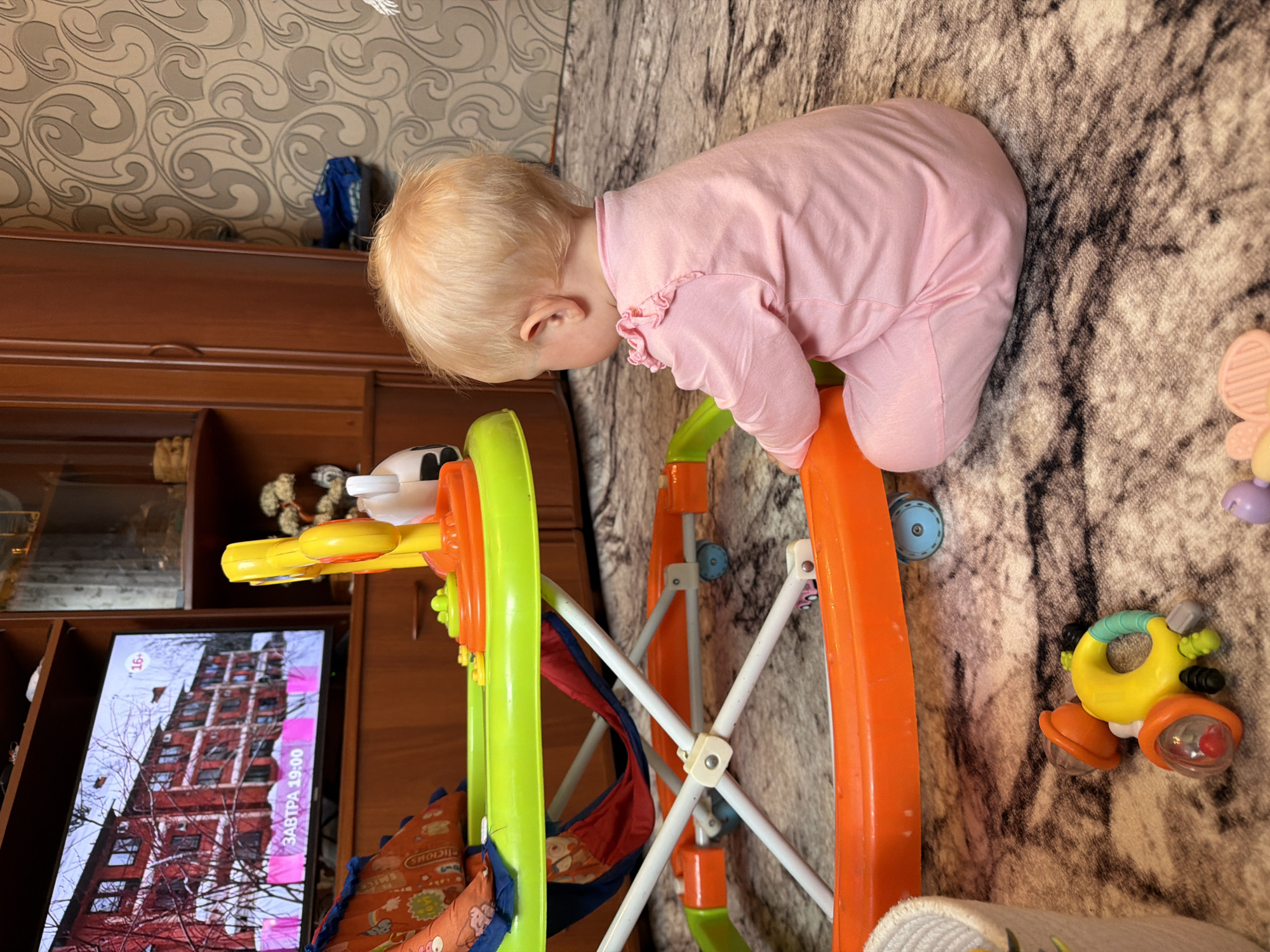 A child in a pink outfit sits on a rug facing a television, with a baby walker in front of them. The television displays an image of a building with the text "ЗАВТРА 19:00" which translates to "TOMORROW 19:00". A child's toy rattle is on the rug. The background includes a wooden cabinet and patterned wallpaper. The scene appears to be an indoor domestic setting, possibly in a living room. The provided location of Sovkhoza "Razdol'e", Russia, is a rural settlement. Without additional visual cues specific to this location, it is not possible to confirm its presence within the image.