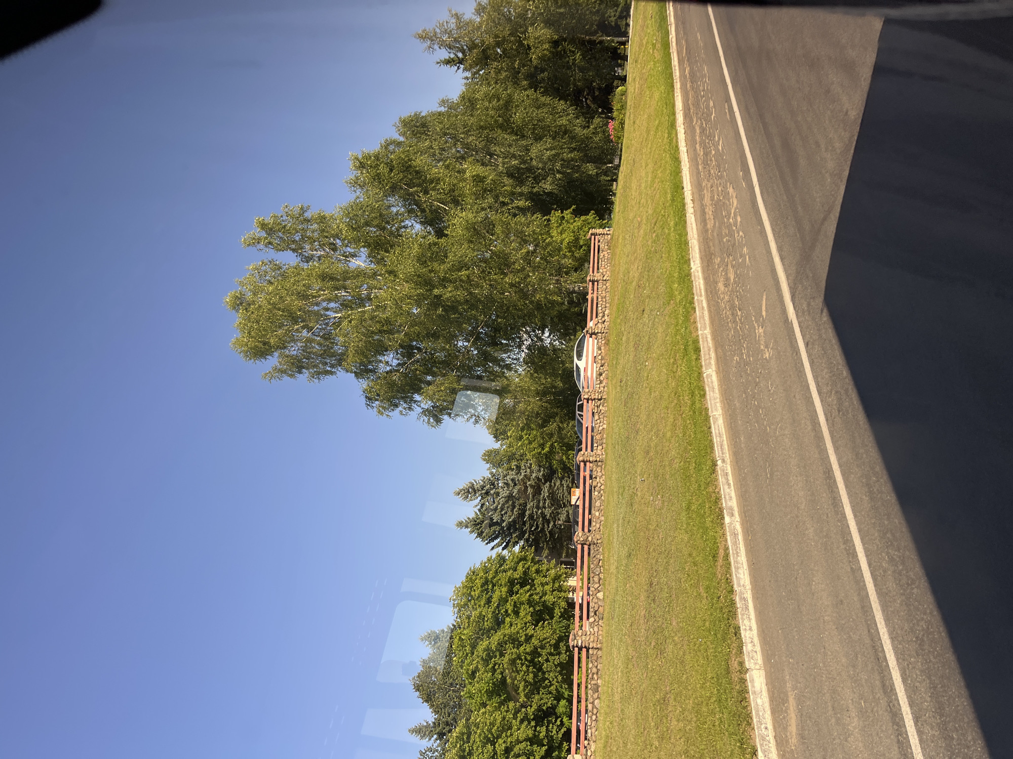 The image depicts a roadside scene with a clear blue sky overhead. In the mid-ground, a low stone and wood fence runs horizontally across the frame, separating a grassy embankment from an area with trees and some parked cars. The trees are a mix of deciduous and coniferous varieties, with large, leafy deciduous trees dominating the right side of the image, and several darker green coniferous trees visible towards the left. A white car with a red stripe is parked on the other side of the fence. The foreground shows a paved road with a white line marking the lane. The overall impression is that of a quiet, sunny day in a semi-rural or suburban area. The location is identified as being in the city of Myadzyel, Belarus.