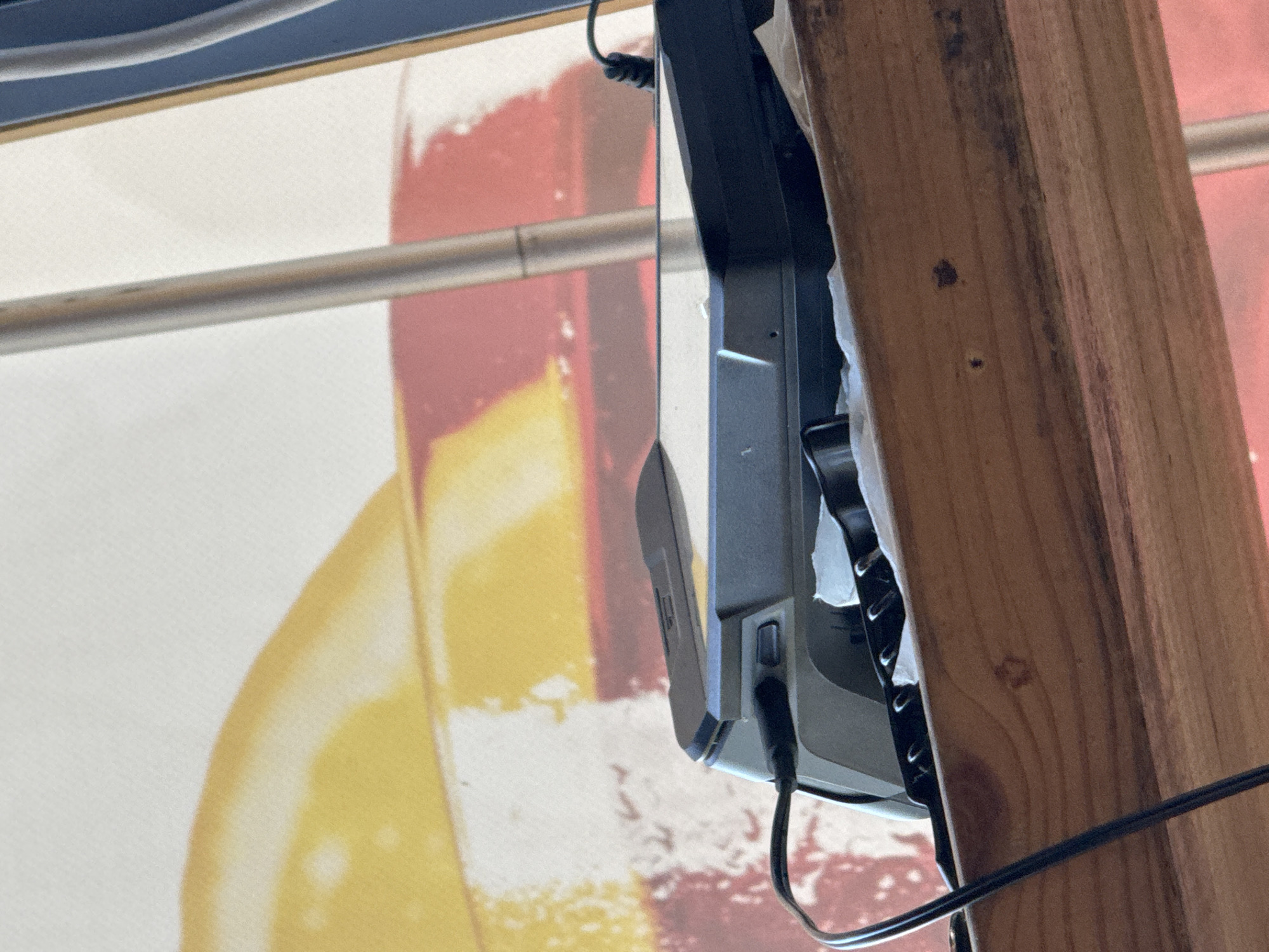 A close-up, indoor shot shows a black electronic device with a partially open lid resting on a wooden surface. The device has visible ports and is connected to a black cable. The background features a large, abstract print with warm colors like yellow and red, possibly a mural or a large piece of art. The scene is likely within an establishment in Burgas, Bulgaria, given the context of the prompt.