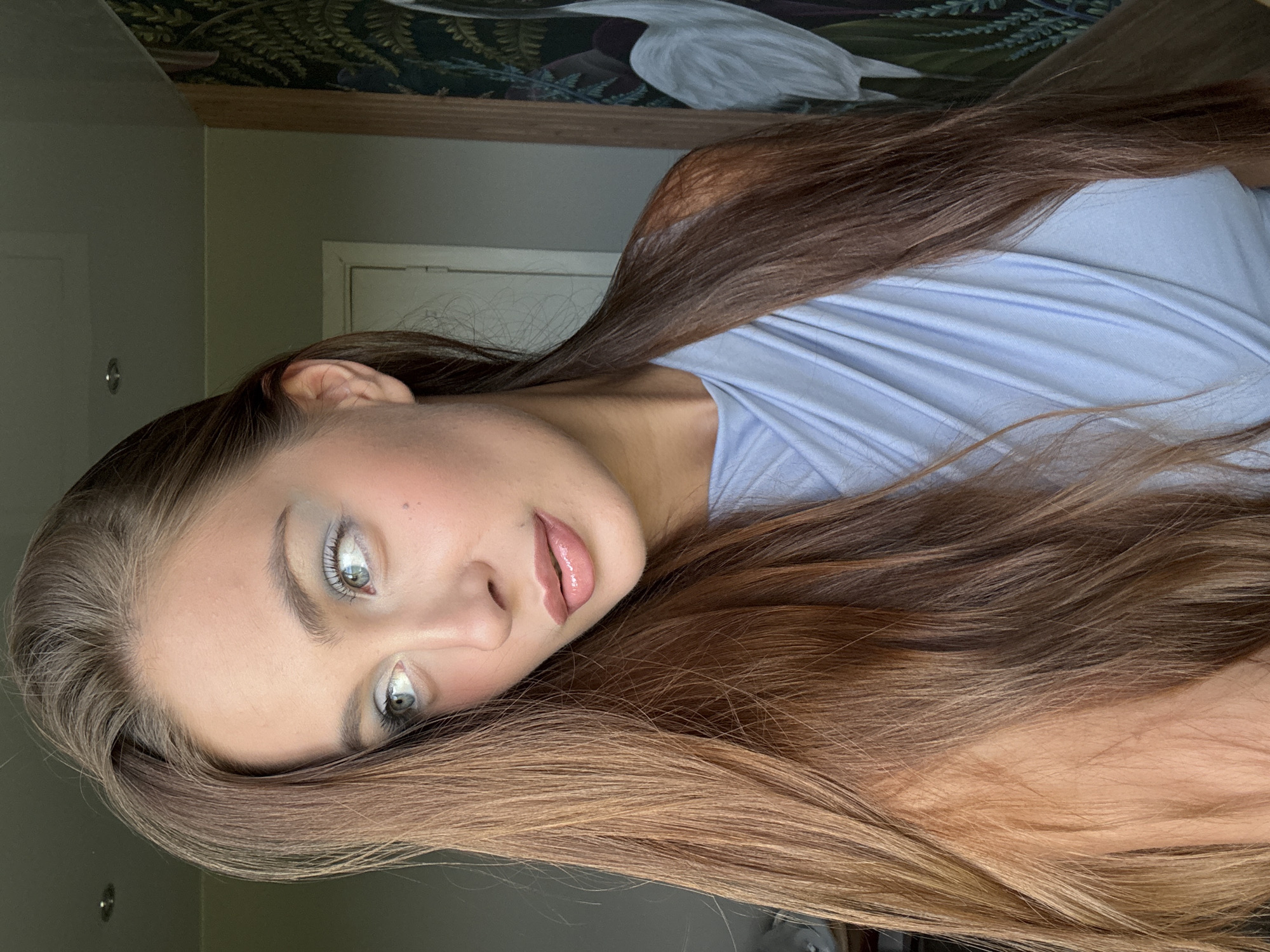 This media file features a close-up portrait of a young woman with long, brown hair. Her head is tilted, and her gaze is directed slightly away from the camera, giving her a contemplative or perhaps shy expression. She is wearing a light blue garment. Her makeup includes subtle eyeshadow and lip gloss, enhancing her natural features. The background appears to be indoors, with a wall and what looks like a door or closet. A decorative element, possibly wallpaper or artwork, is visible along the top edge, featuring a depiction of foliage and a white bird. The lighting suggests it might be daytime, possibly near a window, as there are shadows and highlights on her skin. No visible text is present in the image. The image is oriented with the woman's head on the left side, facing right, and her body extending towards the right.