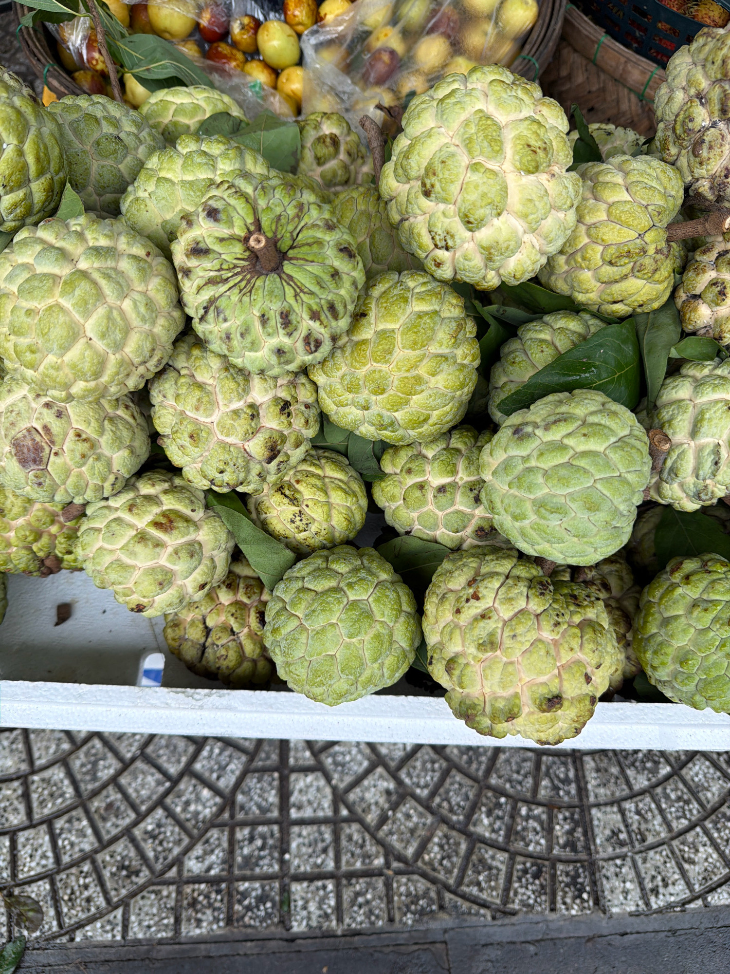 A close-up, overhead shot captures a pile of ripe custard apples, also known as "na" in Vietnamese, displayed for sale at an outdoor market stall in Da Nang, Vietnam. The fruits are characterized by their light green, bumpy skin and are nestled amongst green leaves. In the background, other fruits, such as yellow apples and possibly rambutan, are visible in baskets and plastic bags. The market stall appears to be set up on a street with a patterned pavement, suggesting a typical urban market scene in Da Nang. The overall impression is one of abundance and freshness, highlighting a common sight in Vietnamese fruit markets.