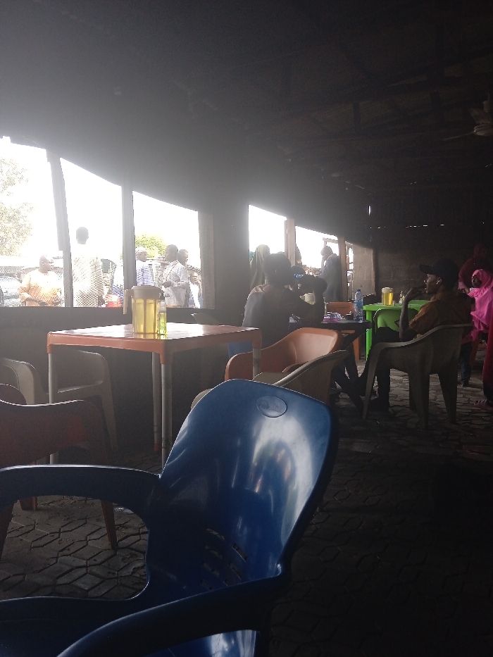 A dim interior scene shows an eatery with plastic chairs and tables. Several people are visible; some stand by the windows, while others sit at the tables. A man in a cap sits in a chair, and a woman in a pink outfit stands nearby. Bottles and pitchers are placed on the tables. The floor is paved with a patterned surface. The background is partially obscured by bright sunlight streaming through the windows.