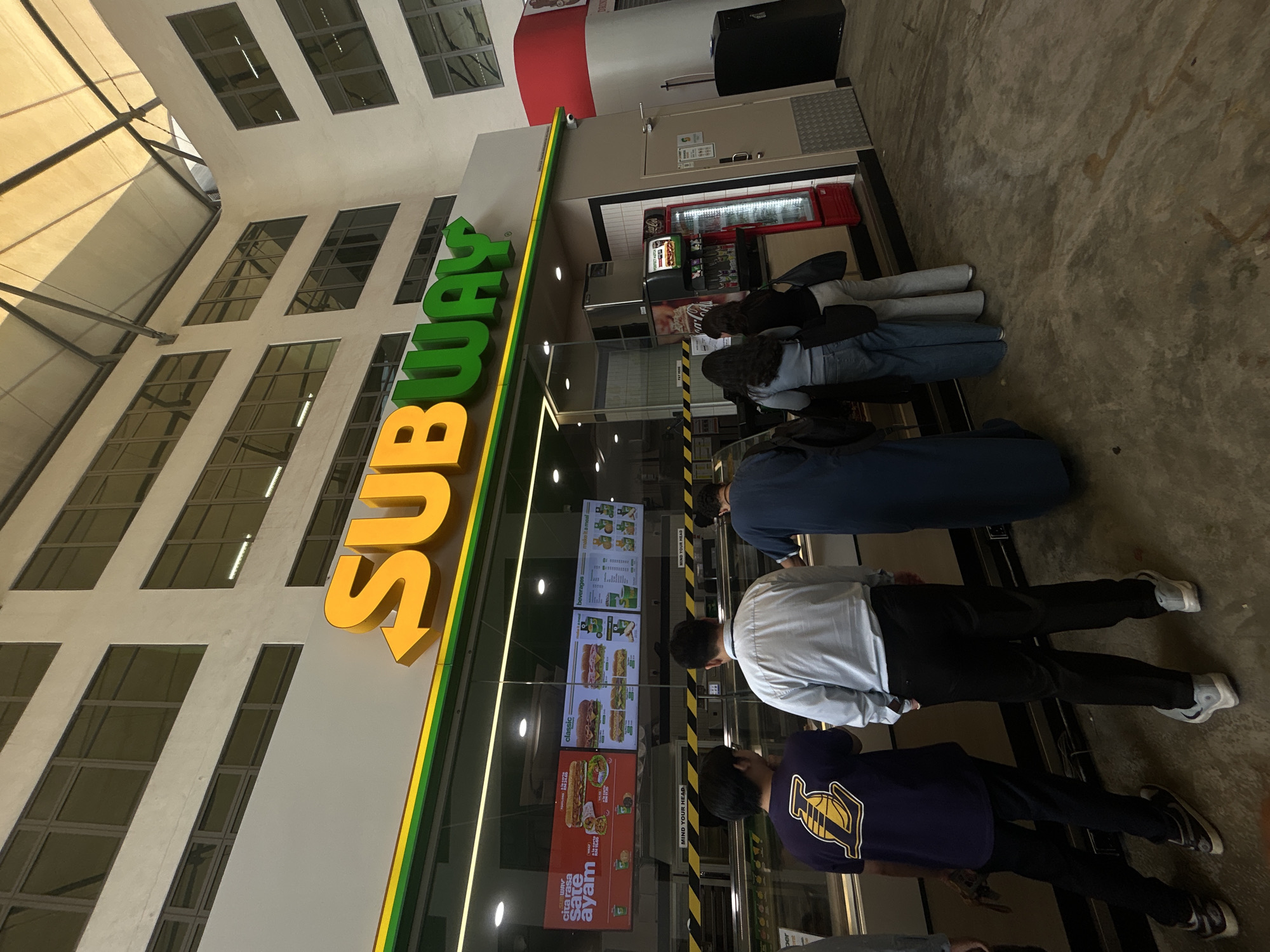 The image shows the exterior of a Subway restaurant. People are waiting in line inside the restaurant. Menu boards are visible. The sign for Subway is displayed prominently on the building. The exterior architecture features multiple windows. The location appears to be in Kuala Lumpur, Malaysia, based on the signage in Malay language on the menu board.