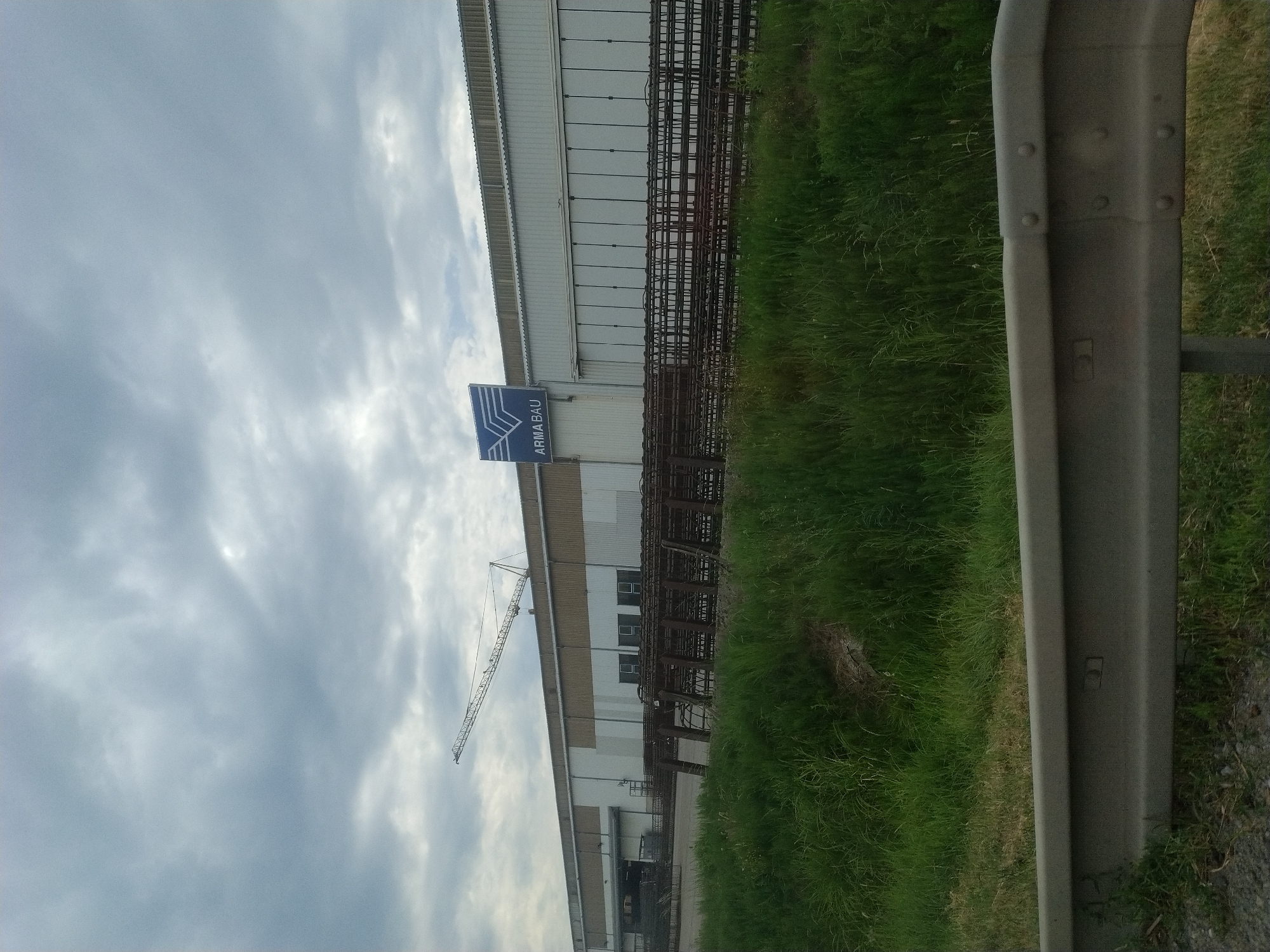 A long industrial building with a corrugated metal roof and white siding stands under a cloudy sky. A crane is visible on the left side of the building. A blue sign with the logo "ARMABAU" is mounted on the facade. In front of the building, piles of rebar are stacked. Lush green grass and weeds grow in front of the rebar piles and extend to a gray metal guardrail in the foreground. The location is the ARMA-BAU company in Kaplice, Czechia.