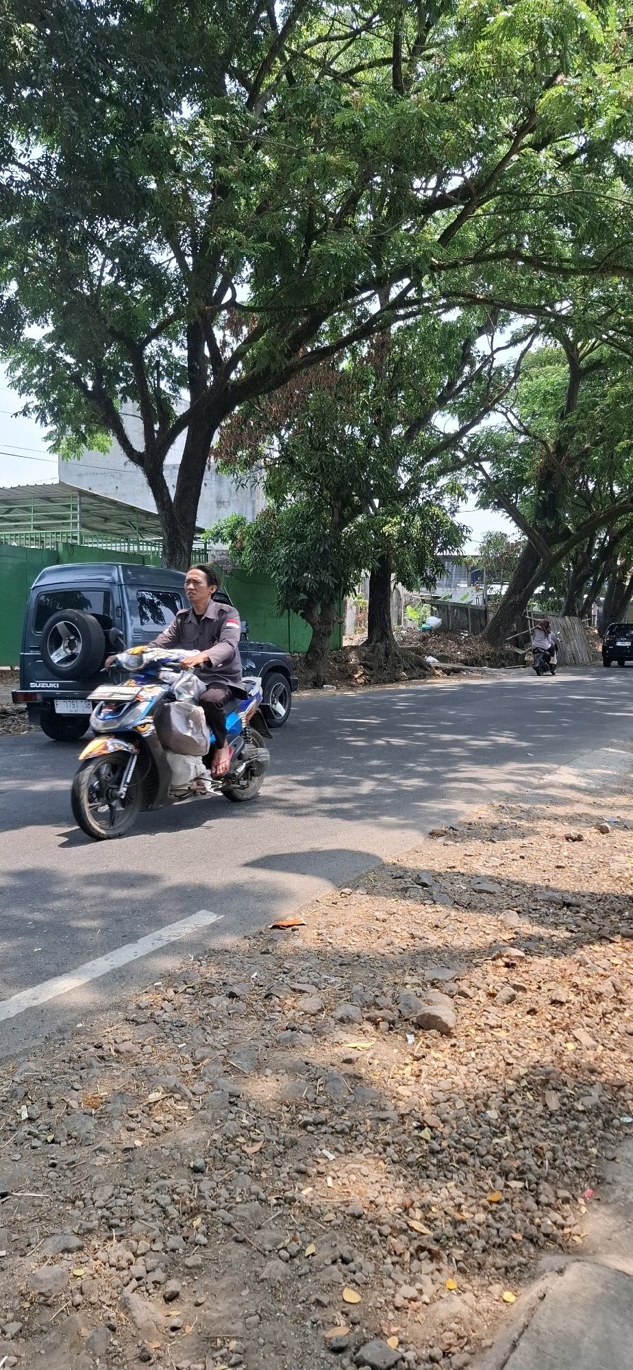 An Indonesian man wearing a dark uniform with a red and white stripe on his sleeve rides a scooter on a paved road in Susukan, Indonesia. He is on the left side of the road, heading towards the viewer. Behind him, on the left, is a dark green Suzuki Jimny with a spare tire mounted on the back. The road is lined with large trees with dense foliage, casting dappled shadows on the asphalt. The ground beside the road is rough and covered with gravel, leaves, and some litter. In the background, there are buildings and more trees. Another scooter is visible further down the road on the right, with a person on it facing away from the camera.