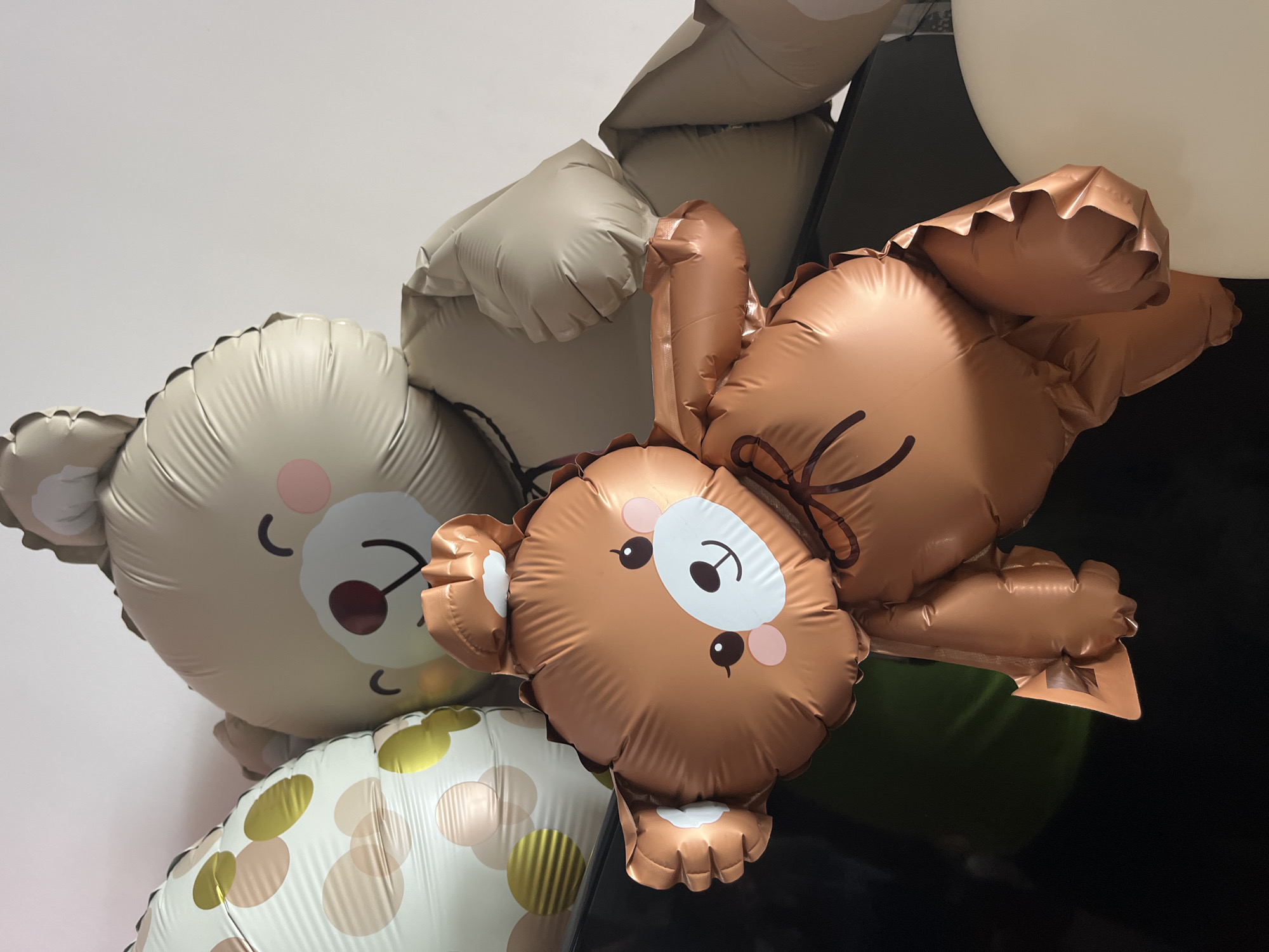 The image shows several balloons, including two bear-shaped balloons. One bear balloon is tan with a simplified face design. Another bear balloon is brown and has a slightly different face design. Other balloons in the composition include a white balloon with gold and tan polka dots, a beige balloon, a green balloon, and a white balloon. The background is a mix of white wall and dark, possibly black, furniture. It is not possible to confirm any location.